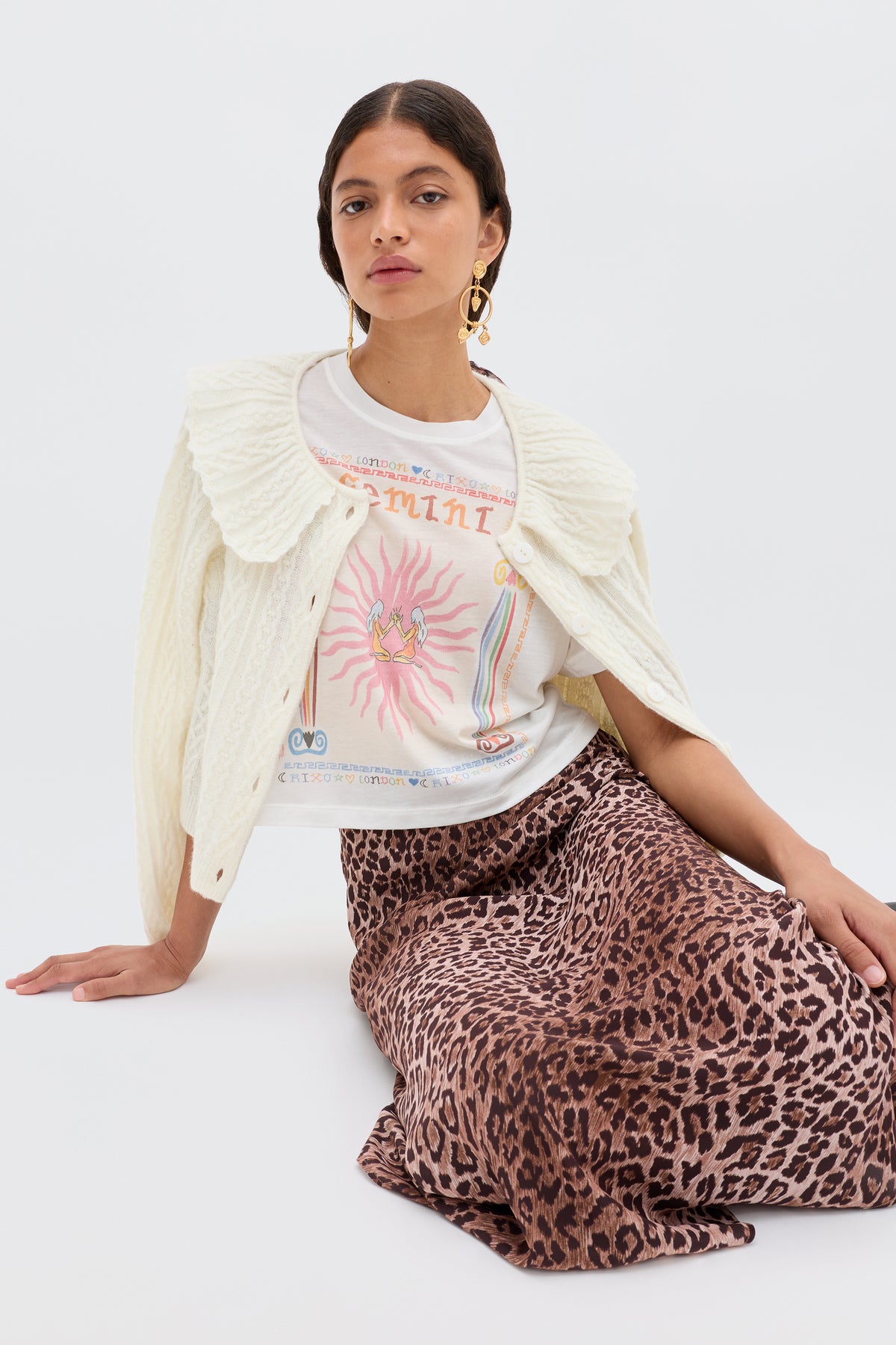 A woman sits on the floor, wearing the Ria - Gemini T-shirt under a white knit sweater, paired with a leopard print skirt and large gold earrings, set against a plain light background.