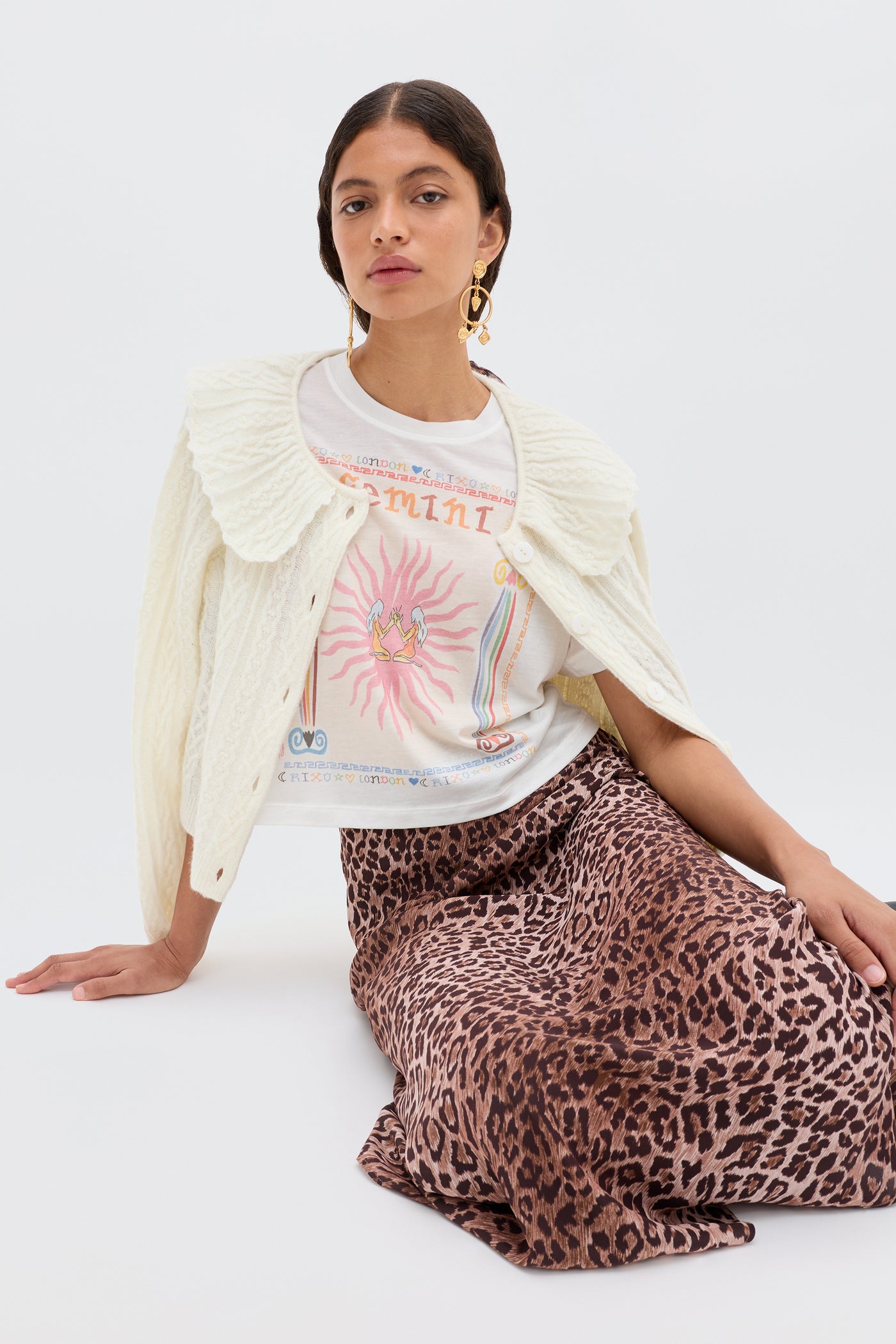 A woman sits on the floor, wearing the Ria - Gemini T-shirt under a white knit sweater, paired with a leopard print skirt and large gold earrings, set against a plain light background.