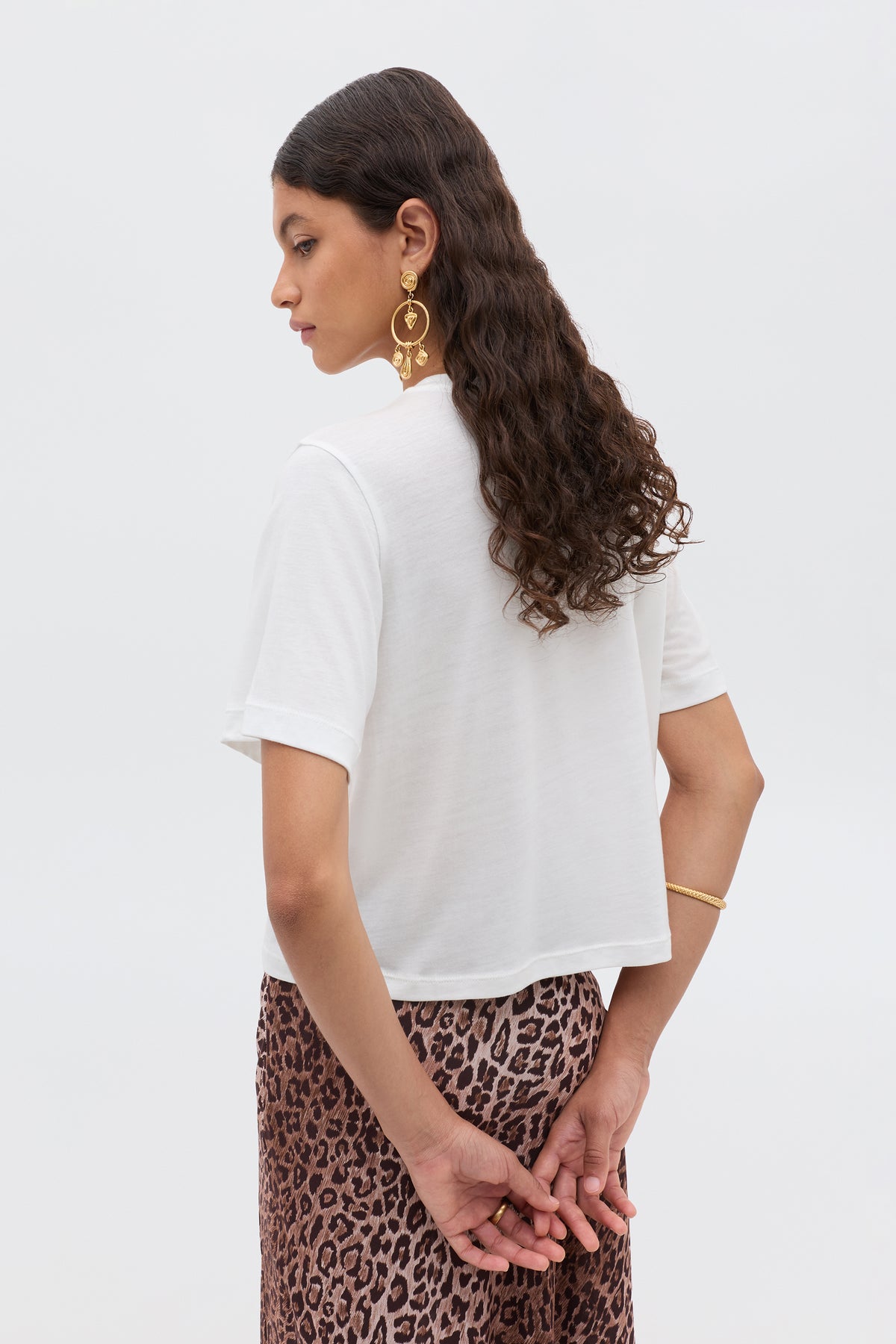 A woman with long wavy hair, hands clasped behind her back, wears a white sheer Ria - Gemini T-shirt and a leopard print skirt, accessorizing with gold earrings and a bracelet.