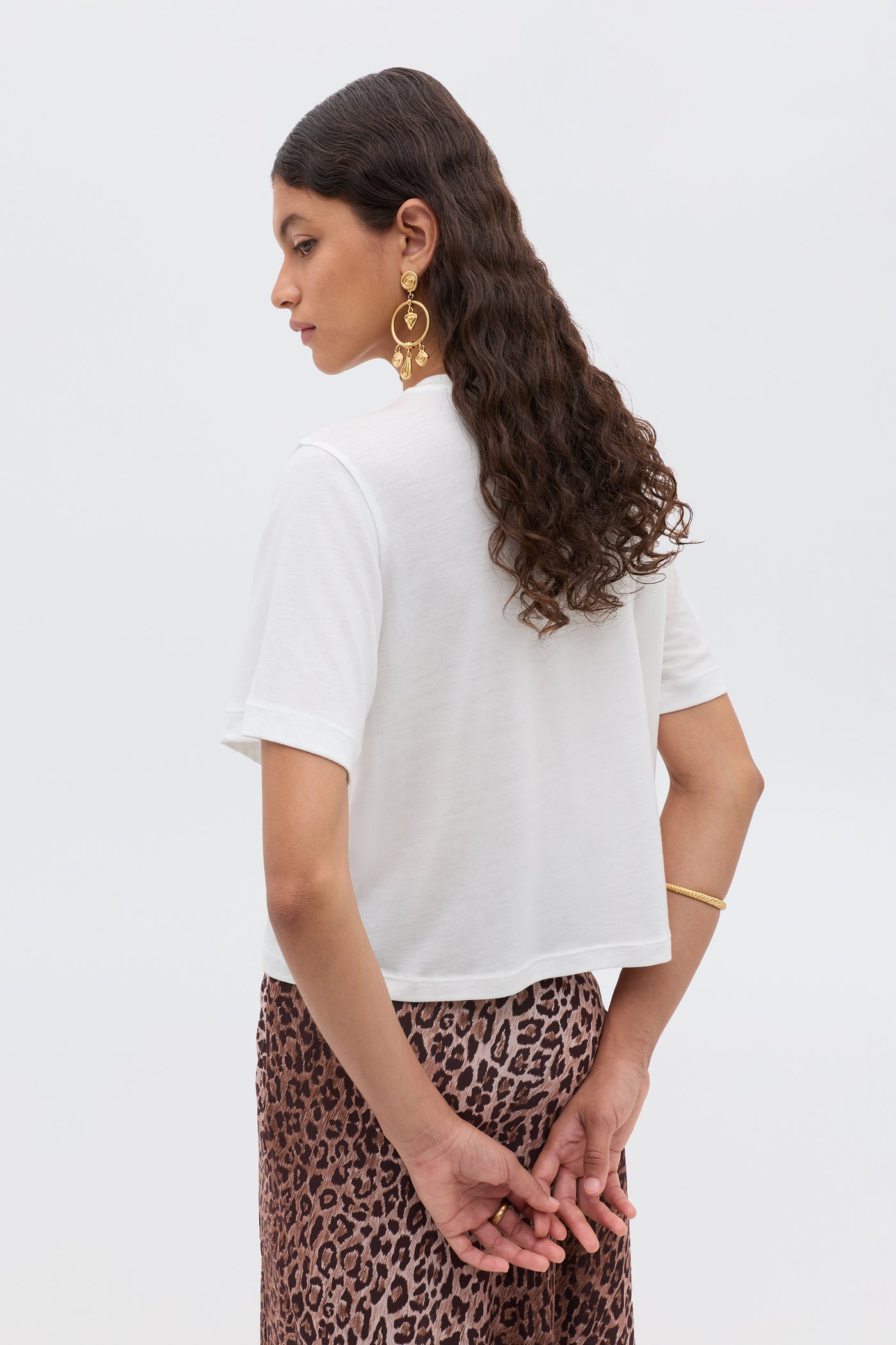 A woman with long wavy hair, hands clasped behind her back, wears a white sheer Ria - Gemini T-shirt and a leopard print skirt, accessorizing with gold earrings and a bracelet.