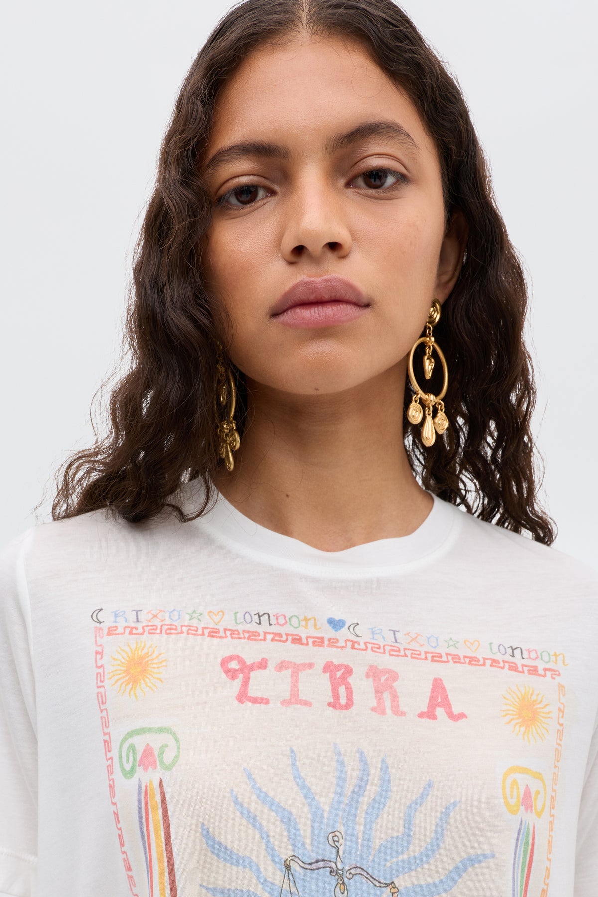 A woman with long, wavy brown hair wears Ria - Libra: gold dangling earrings and a white cropped T-shirt featuring a colorful Libra illustration and astrological symbols. She faces the camera with a neutral expression.