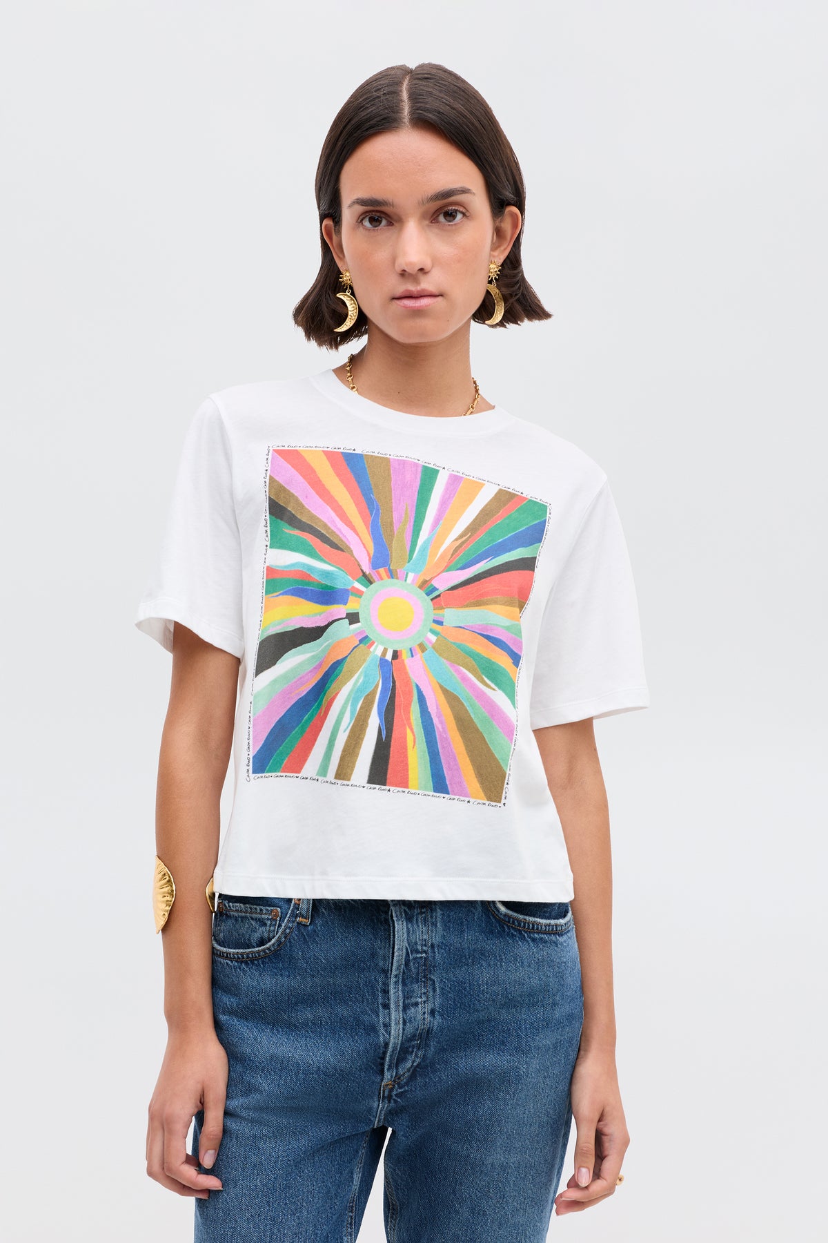 A woman with short brown hair wears the Ria- Casa Soleil Multi T-shirt, featuring a colorful abstract sunburst and cropped hem. She pairs it with blue jeans, large gold earrings, and a bracelet, standing against a light background.