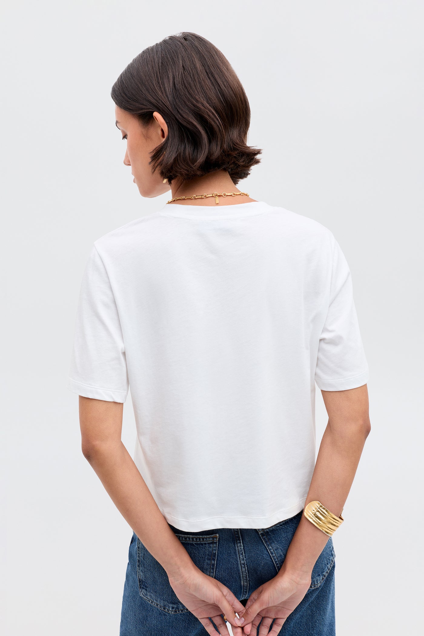 A person with short, dark hair stands with their back to the camera, wearing the Ria- Casa Soleil Multi top and blue jeans. Their hands are clasped behind their back, accessorized with a gold bracelet and necklace.
