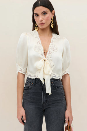 A woman wears the Amara - Champagne, a cream lace short-sleeve blouse with a front bow, paired with high-waisted dark jeans. She has long straight hair, gold earrings, and a neutral expression against a plain background.