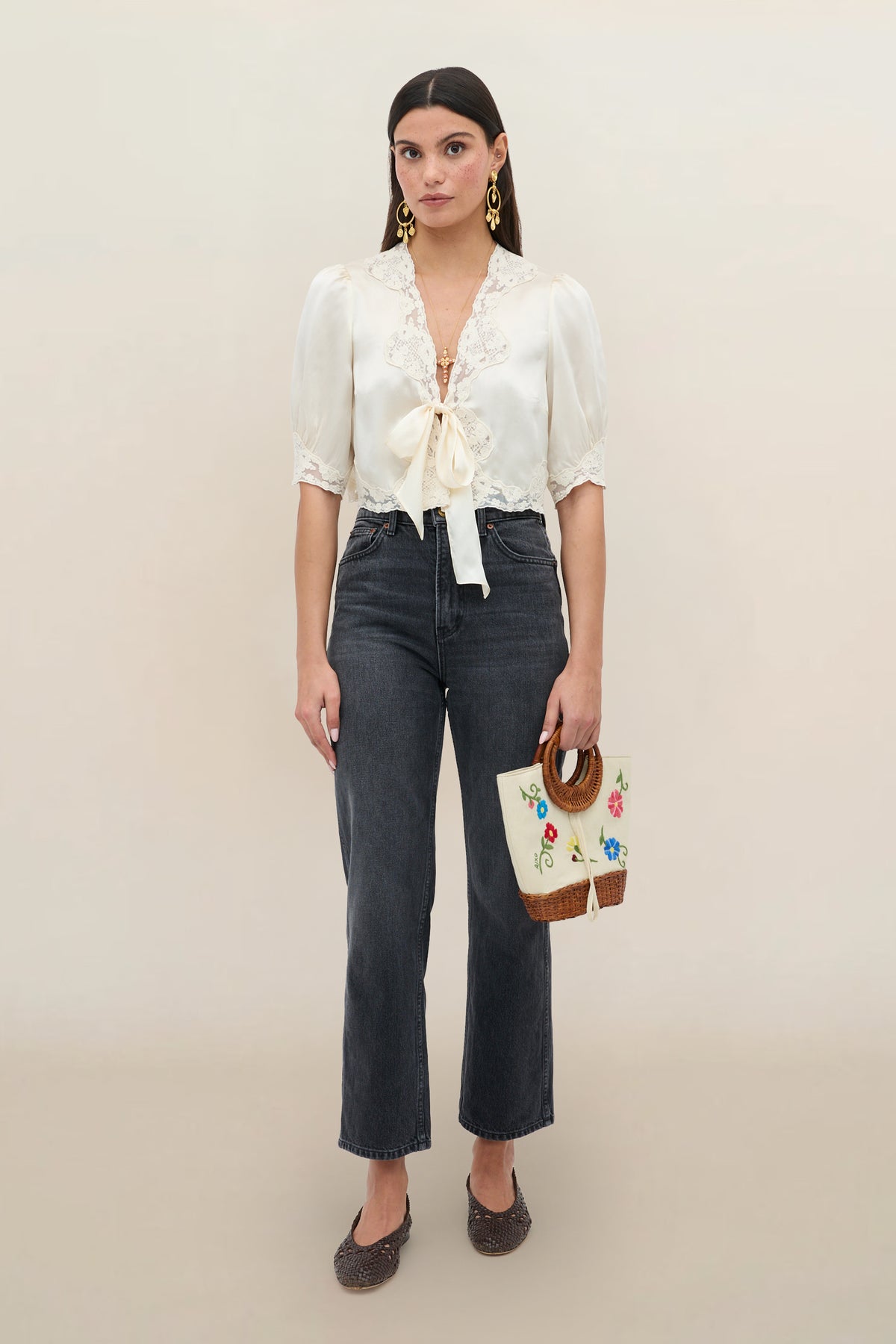 A woman stands against a plain background wearing the Amara - Champagne silk-satin top with lace trim and bow, paired with high-waisted dark jeans, brown shoes, gold earrings, and a woven handbag with embroidered flowers.