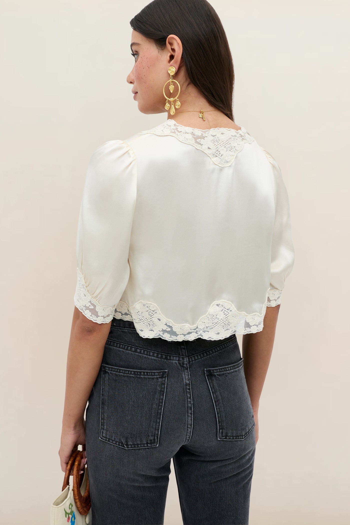 A woman with long dark hair wears the Amara - Champagne, a cream lace-trimmed blouse with puff sleeves, paired with high-waisted dark gray jeans. She holds a small white bag, wears large gold earrings, and faces away from the camera.