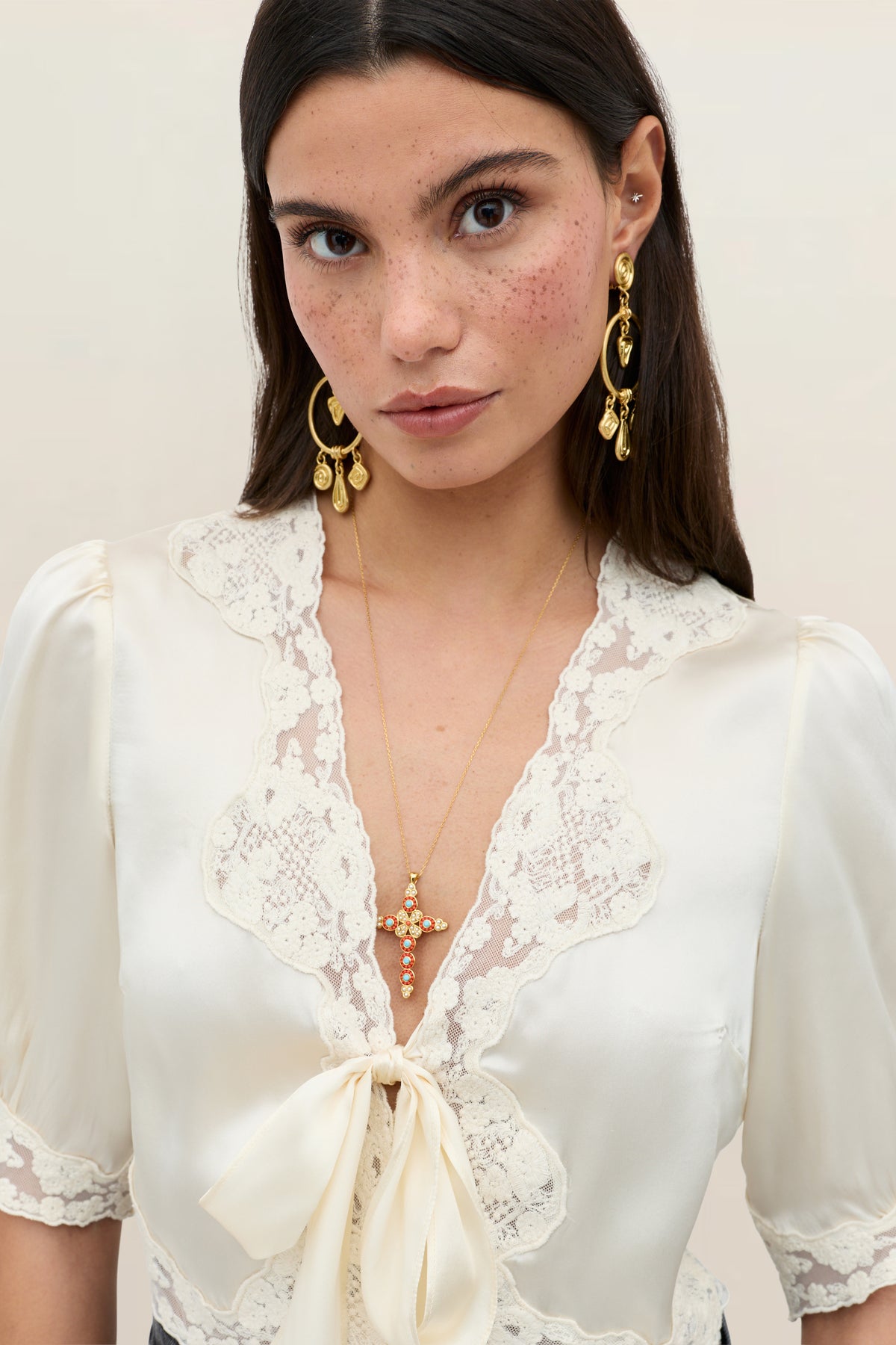 A woman with long brown hair and freckles wears the Amara top in Champagne, which features lace trim and a bow. She accessorizes with gold earrings and a jeweled cross necklace, gazing directly at the camera.
