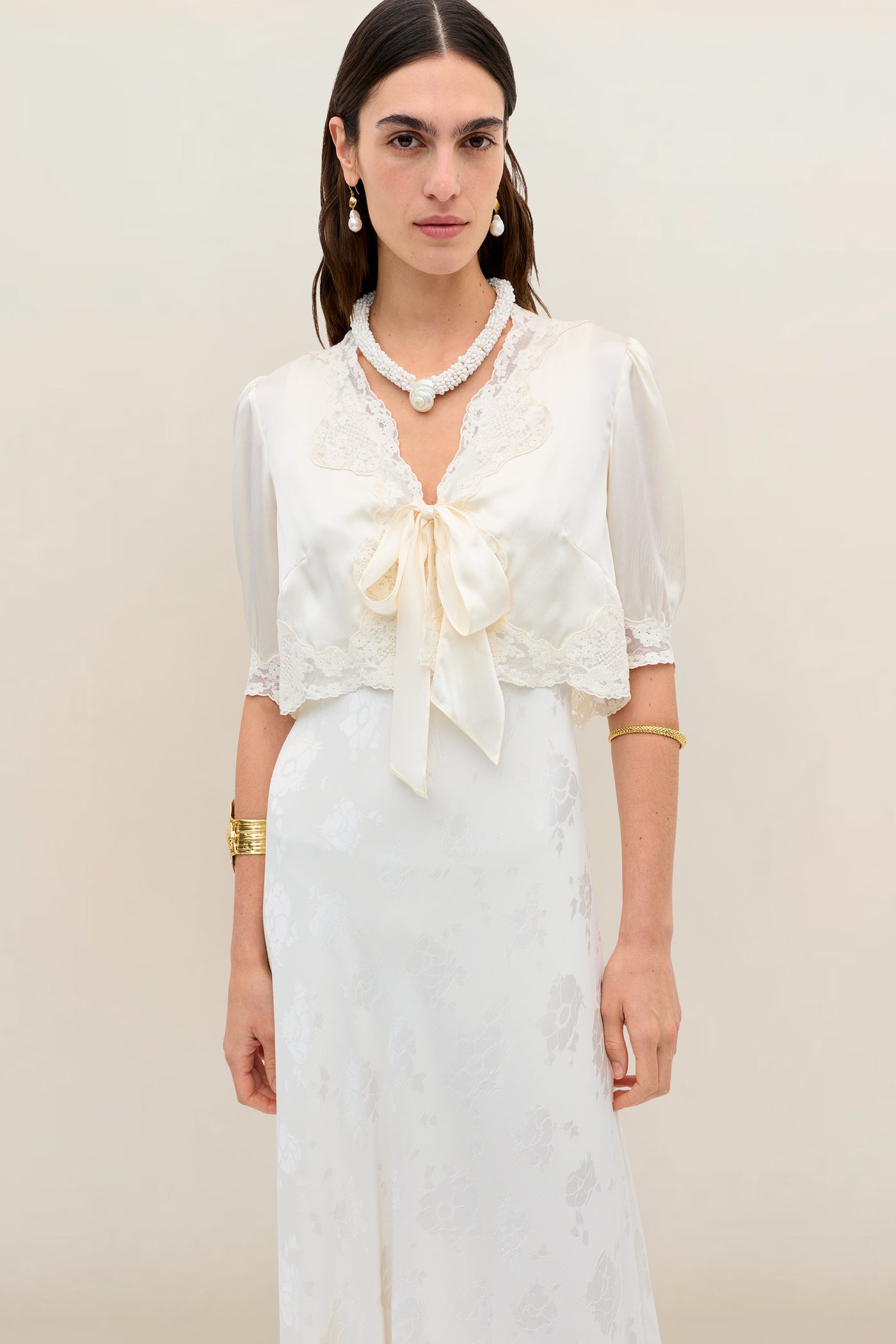 A woman wears the Amara - Champagne, a vintage-style silk-satin cropped jacket with lace trim and a front bow, over a matching long dress, accessorized with gold bracelets, pearl earrings, and a beaded necklace.