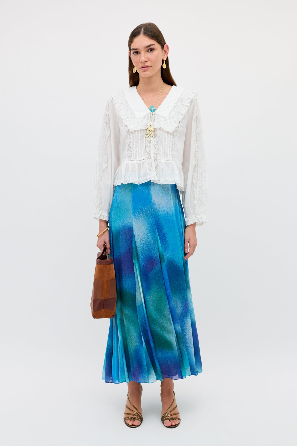A woman stands against a white background wearing the Billy - Jessica Alexandra Blue, a flowing blue and green midi skirt crafted from Liberty fabric—perfect for spring/summer dressing—paired with a long-sleeve white blouse featuring a ruffled collar, strappy sandals, gold earrings, and holding a brown handbag.