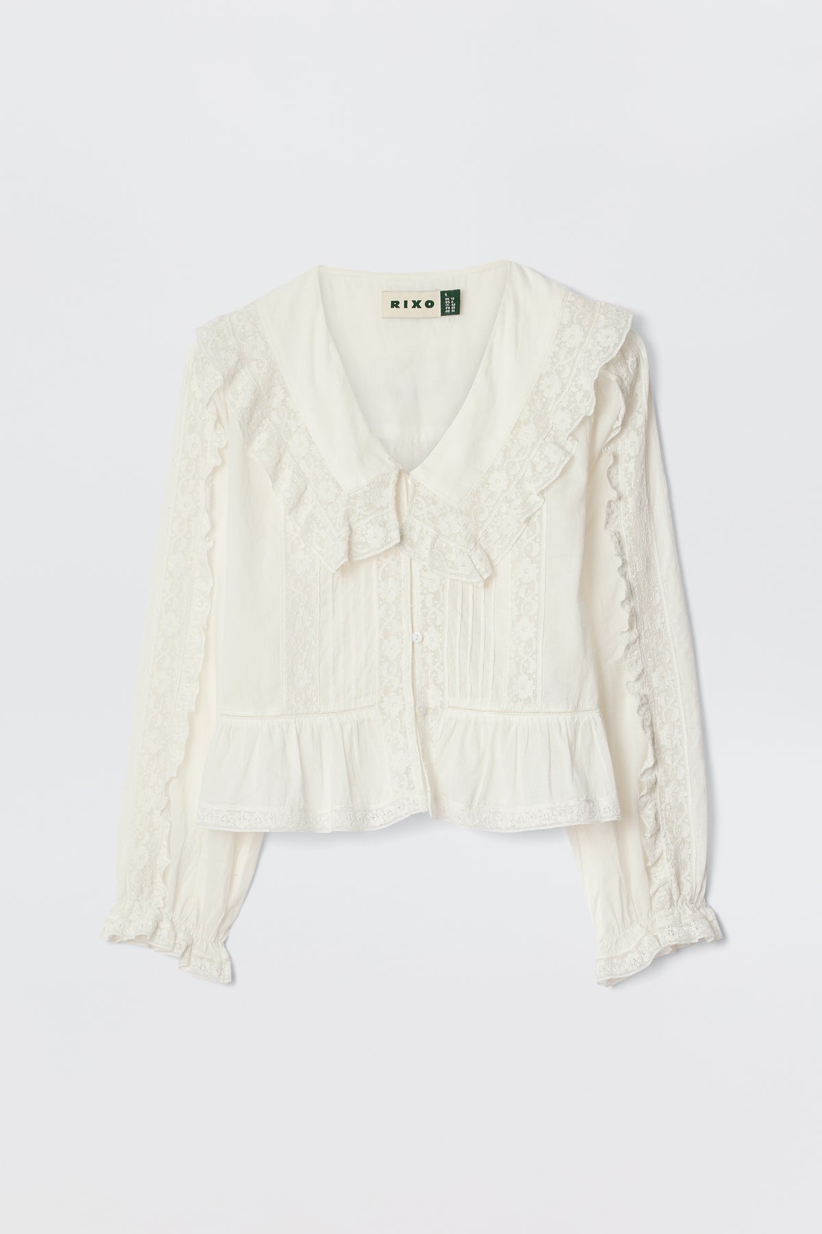 The Maribelle - Ivory is a vintage-inspired, long-sleeved white blouse with lace trim, button-down front, ruffled cuffs, and a wide collar with bow detail, shown on a white background.