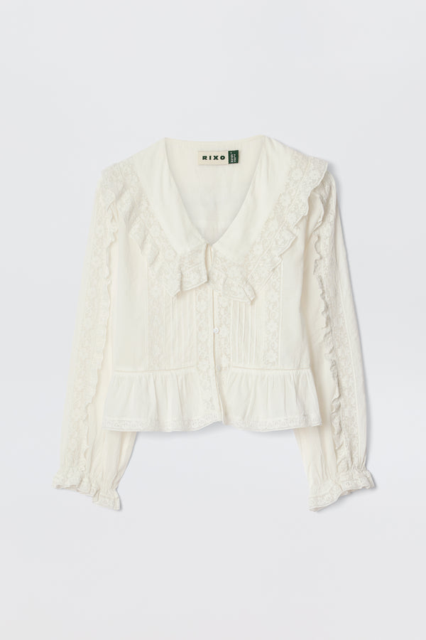 The Maribelle - Ivory is a vintage-inspired, long-sleeved white blouse with lace trim, button-down front, ruffled cuffs, and a wide collar with bow detail, shown on a white background.