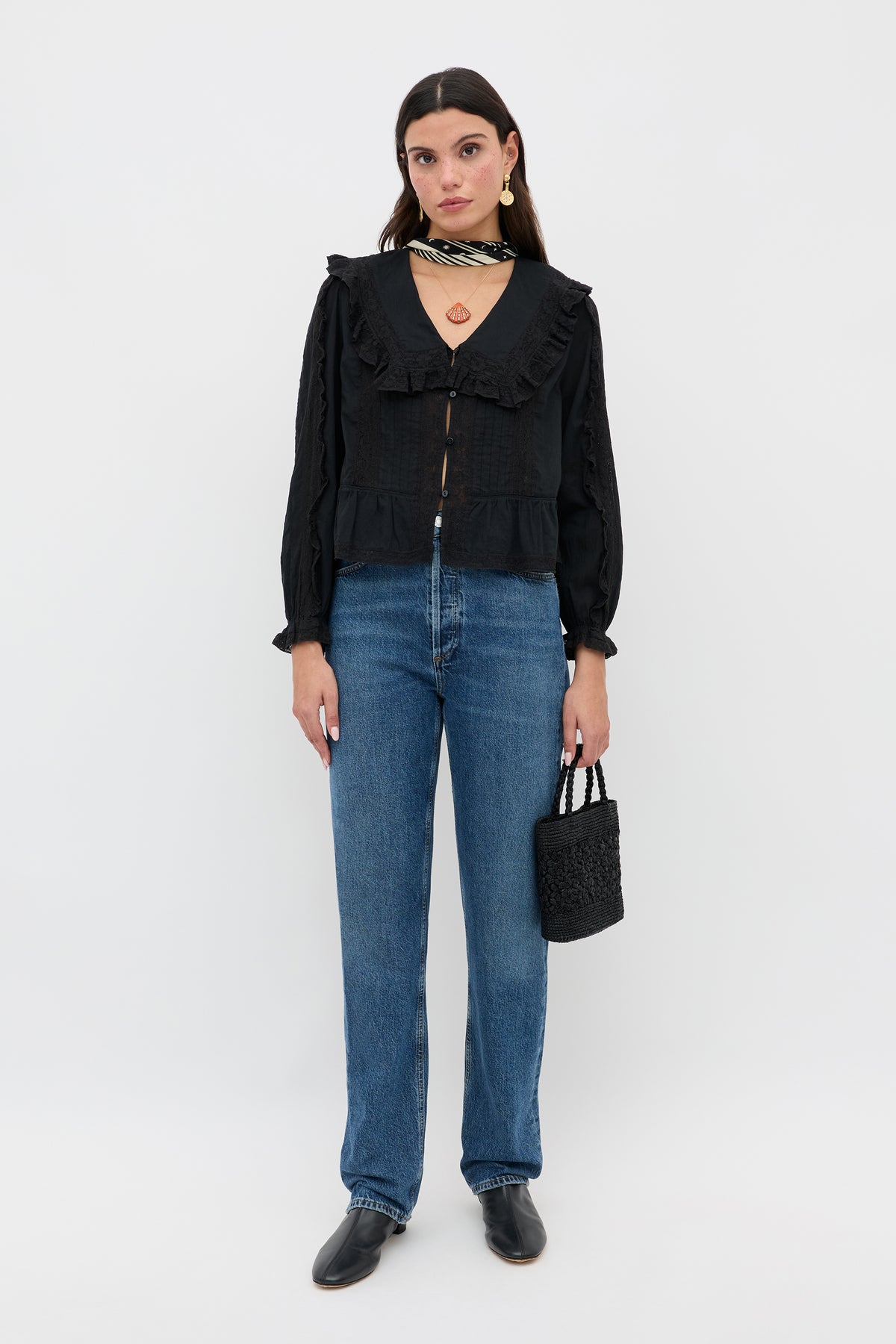 A woman stands against a plain background wearing Maribelle - Black, a vintage-inspired ruffled blouse with a relaxed fit, paired with blue straight-leg jeans, black shoes, a textured handbag, statement jewelry, and long dark hair.