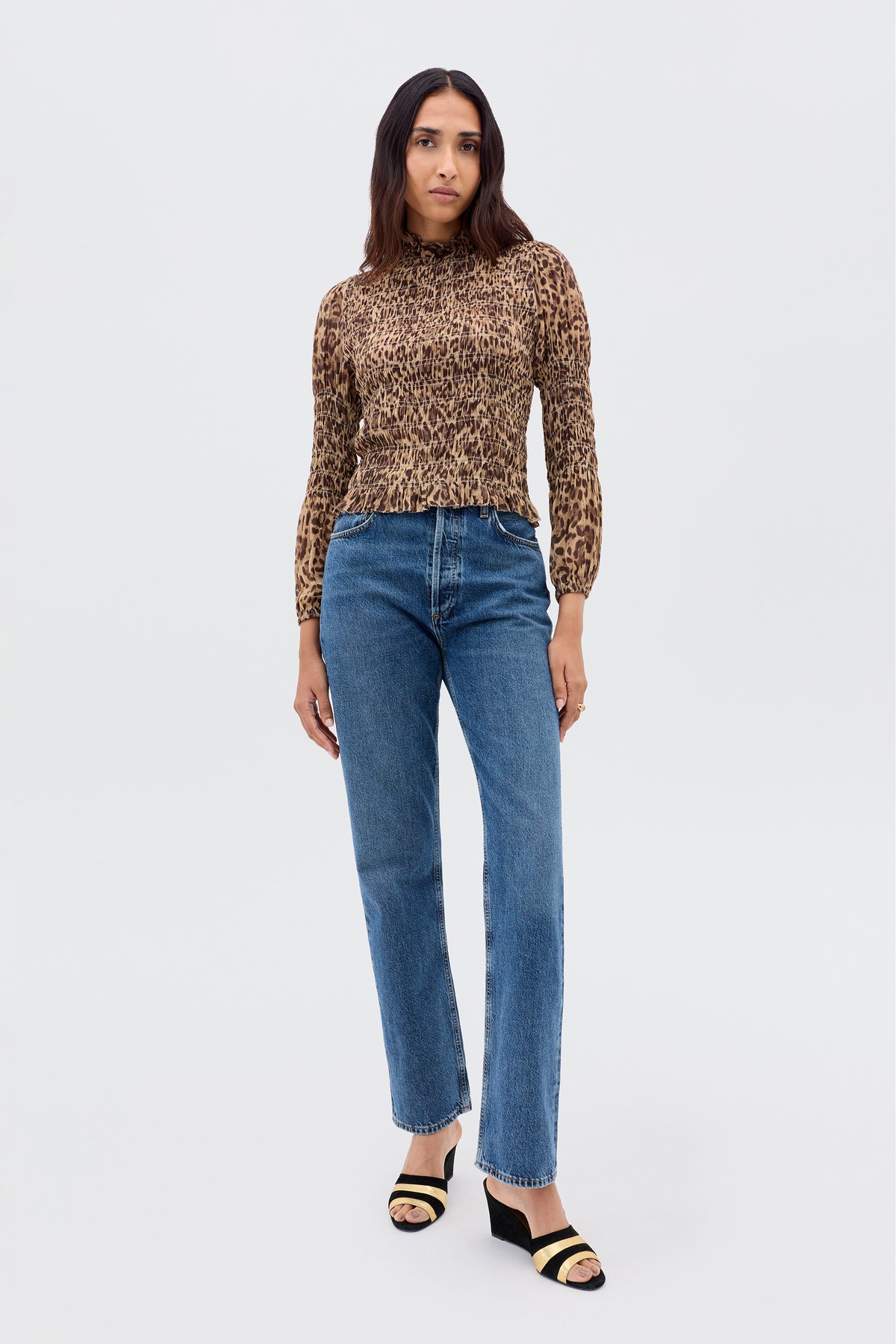 A woman with long, dark hair and a neutral expression wears the Gardenia - Mila Leopard Brown blouse with long sleeves, paired with blue high-waisted jeans and black and beige open-toe heeled sandals against a plain background.