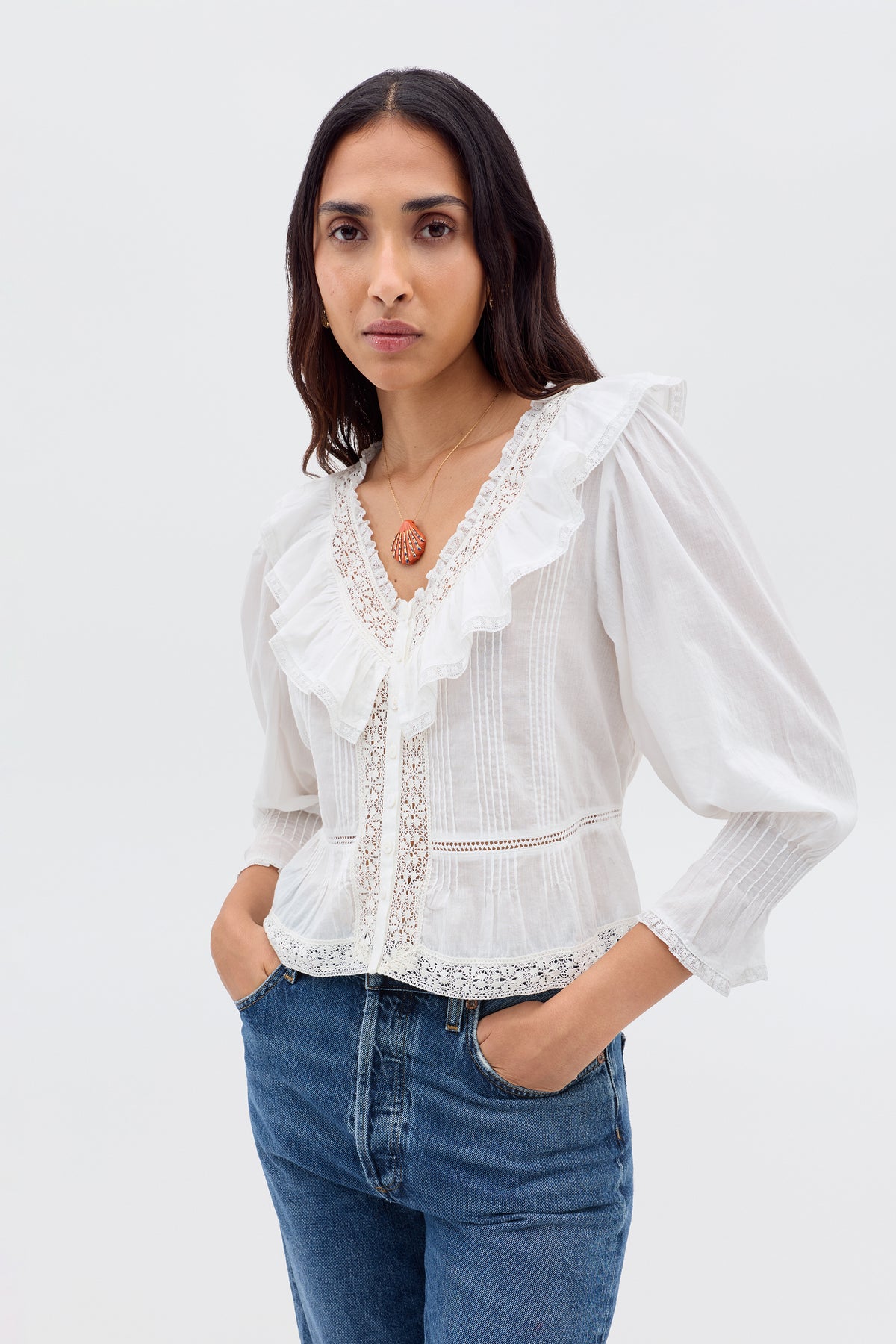 A woman with long brown hair wears the Solene - Ivory blouse—vintage-inspired with puffed sleeves, lace details, and a ruffled V-neckline—paired with blue jeans as she stands against a plain light background, hands in her pockets.
