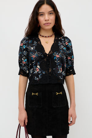 Wearing the “Ursula - Botanical Embroidery Black” blouse, a woman with long dark hair pairs it with a black skirt featuring gold details, accessorizes with a beaded necklace, and carries a red bag as she stands against a plain white background.