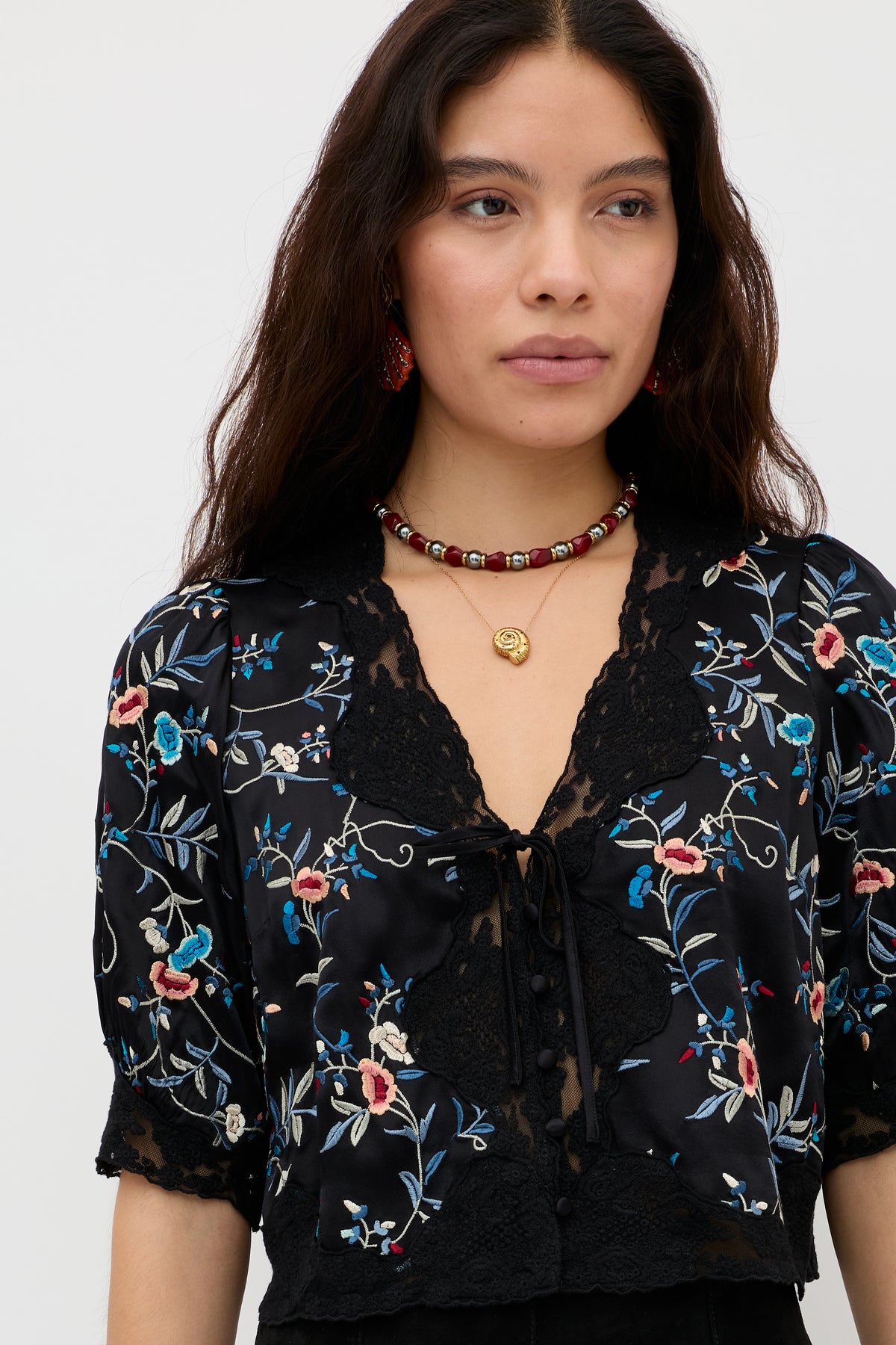 A woman with long dark hair wears the Palmara - Burgundy blouse, featuring colorful floral embroidery. She pairs it with a beaded necklace and drop earrings, standing against a plain light background with a neutral expression.