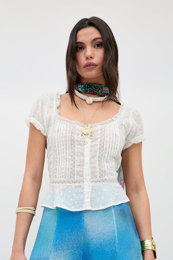 A woman wears the Shereen - Cream sheer eyelet blouse with short puff sleeves, blue ombré pants, layered gold jewelry, and a colorful neck scarf, standing against a light background.