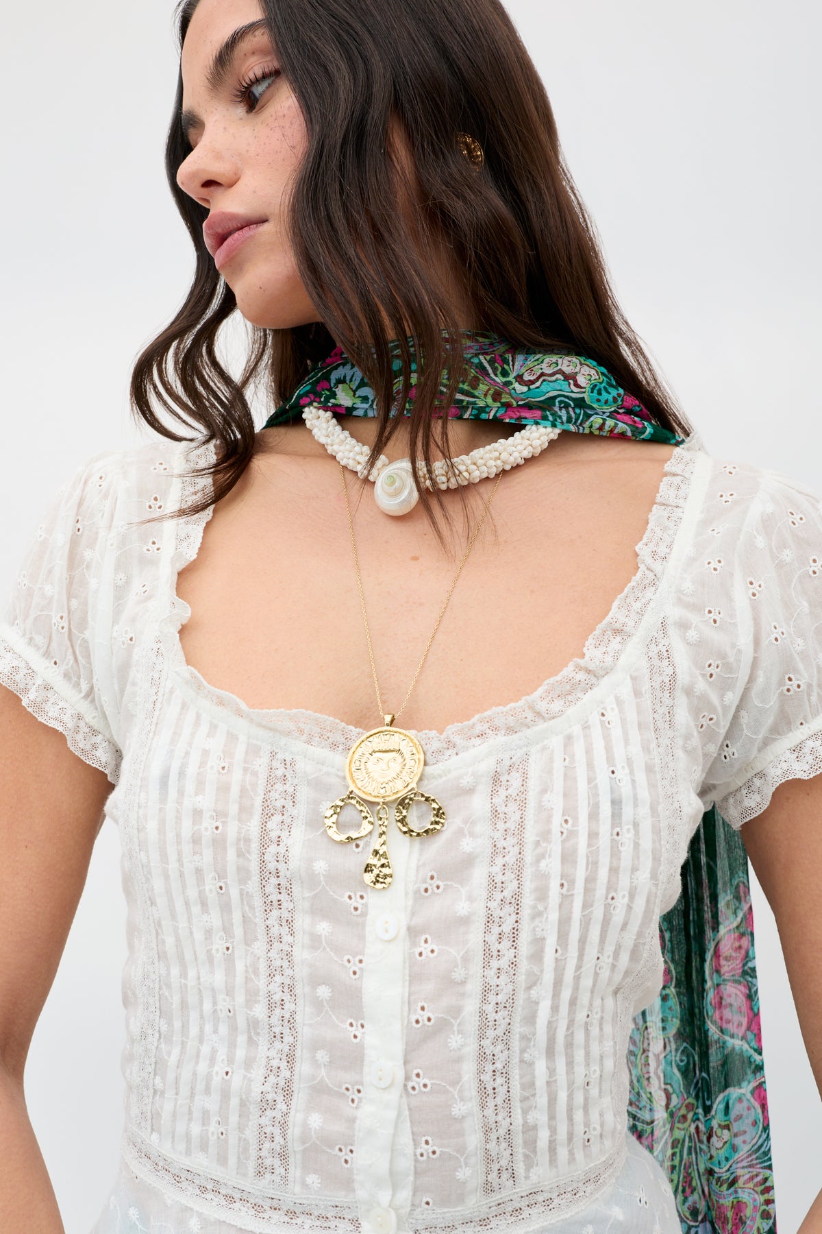 A woman wears the Zahra - Gold layered necklaces with a white lacy button-up blouse and colorful scarf, standing against a plain light background.