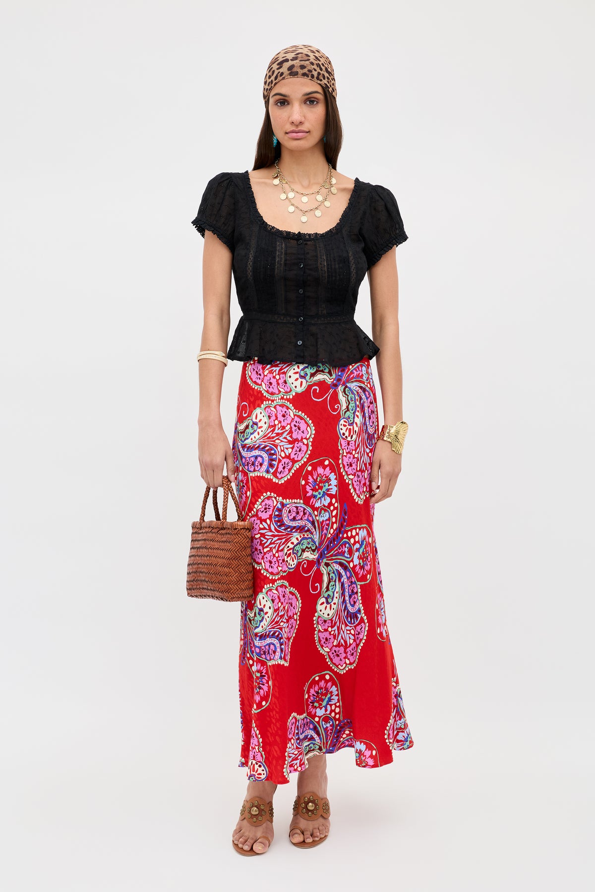 A woman wears the Sandi - Black broderie anglaise cotton blouse with a red paisley skirt, brown sandals, leopard-print headscarf, woven handbag, and layered jewelry, posing against a plain white background.