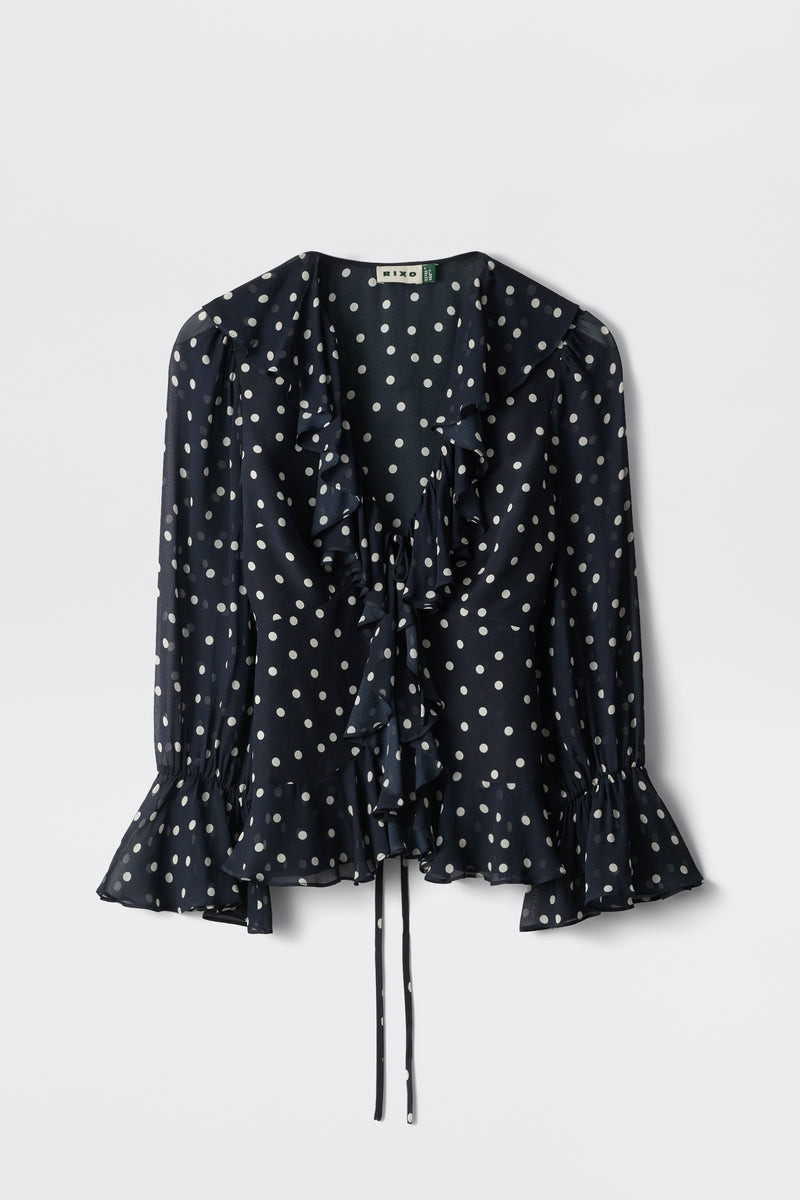 The Ami - Layla Dot Navy is a navy blue long-sleeve blouse featuring white polka dots, a ruffled neckline and cuffs, and a waist tie, shown against a plain white background.
