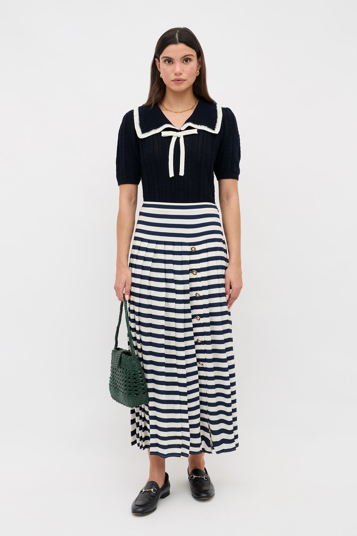 A woman stands against a plain background wearing Pippa - Deep Navy, a short-sleeve sailor-collared top with bow, paired with a high-waisted navy-and-white striped buttoned skirt, black loafers, and holding a green woven handbag.
