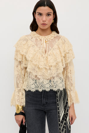 A woman wearing the Faryn - Vintage Cream sheer lace blouse with ruffles, paired with black jeans and a black-and-white patterned scarf hanging from her waist, stands against a plain white background.