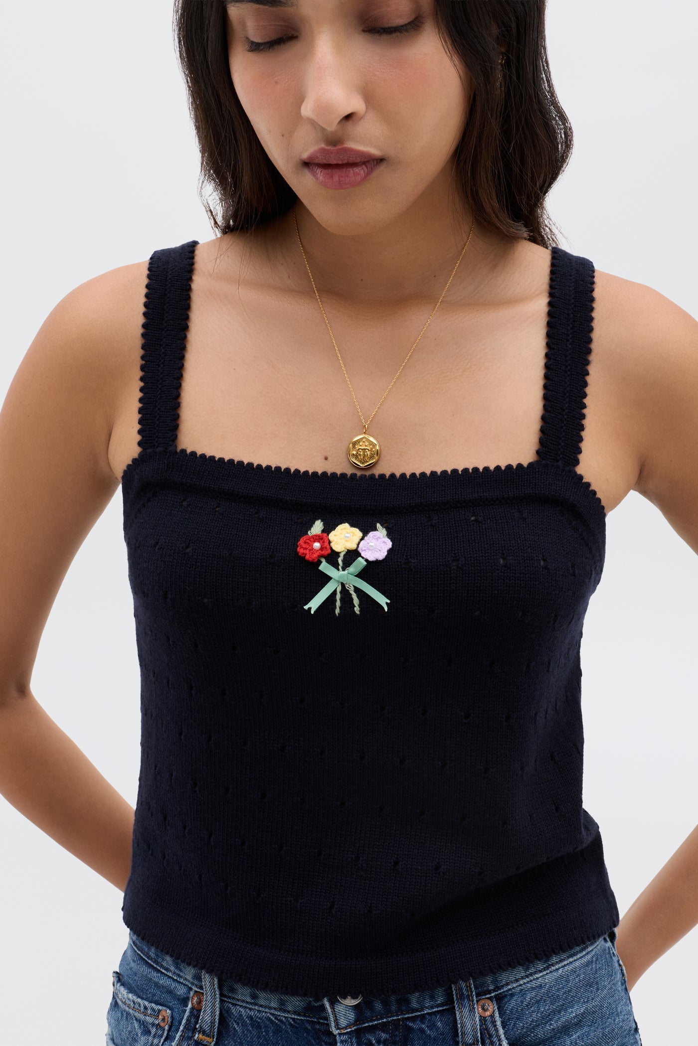A woman in the Perla - Floret Embroidery Deep Navy tank top with a colorful floral motif at the center, gold pendant necklace, long dark hair, and denim jeans stands looking down with arms relaxed at her sides.
