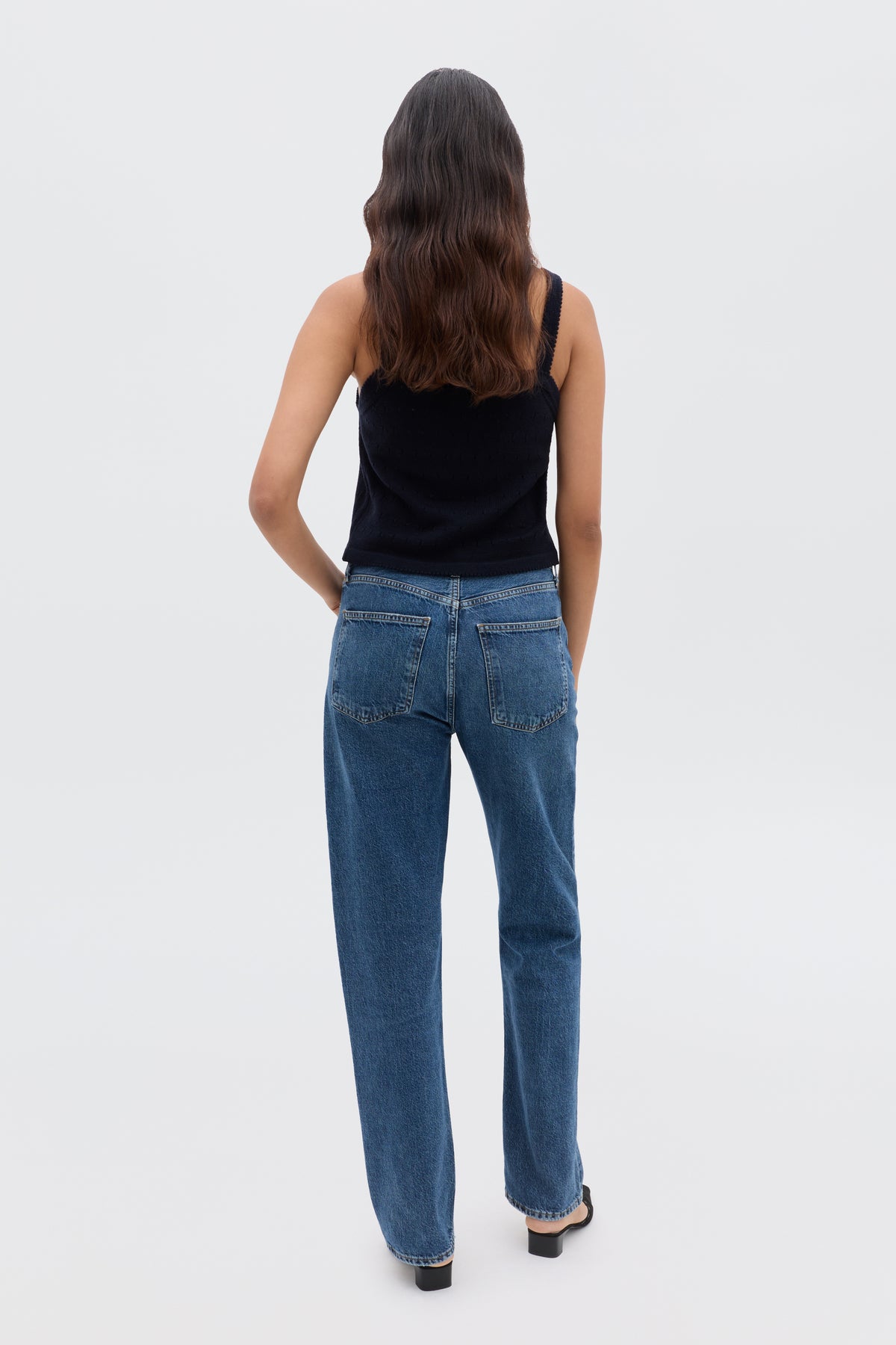 A person with long, wavy brown hair stands facing away, wearing the Perla - Floret Embroidery Deep Navy sweater with blue jeans and black shoes against a plain light background.
