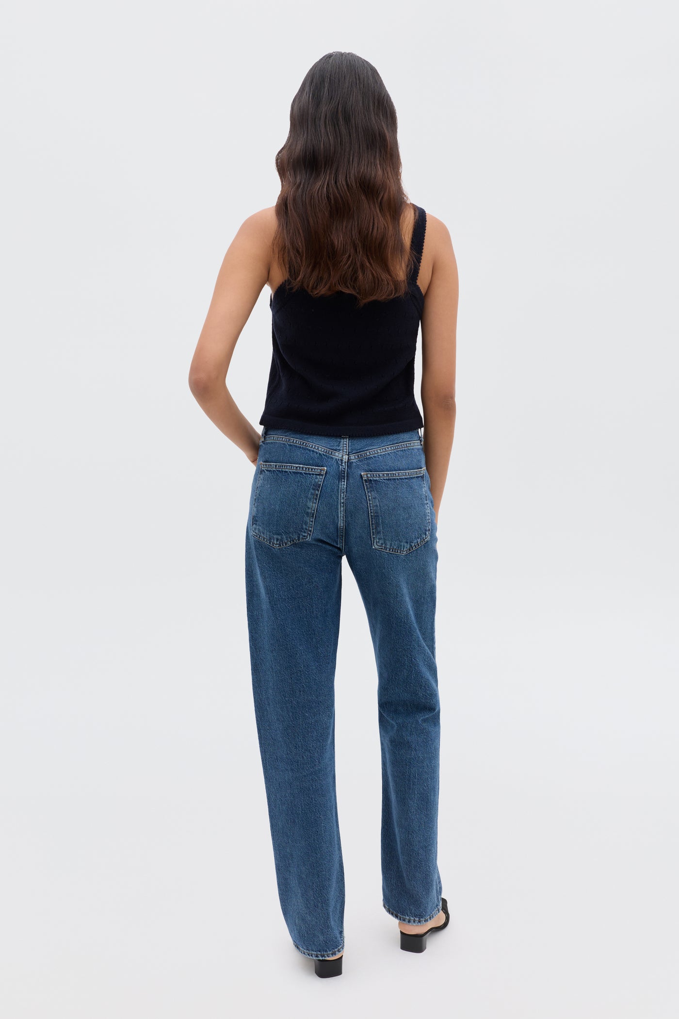 A person with long, wavy brown hair stands facing away, wearing the Perla - Floret Embroidery Deep Navy sweater with blue jeans and black shoes against a plain light background.