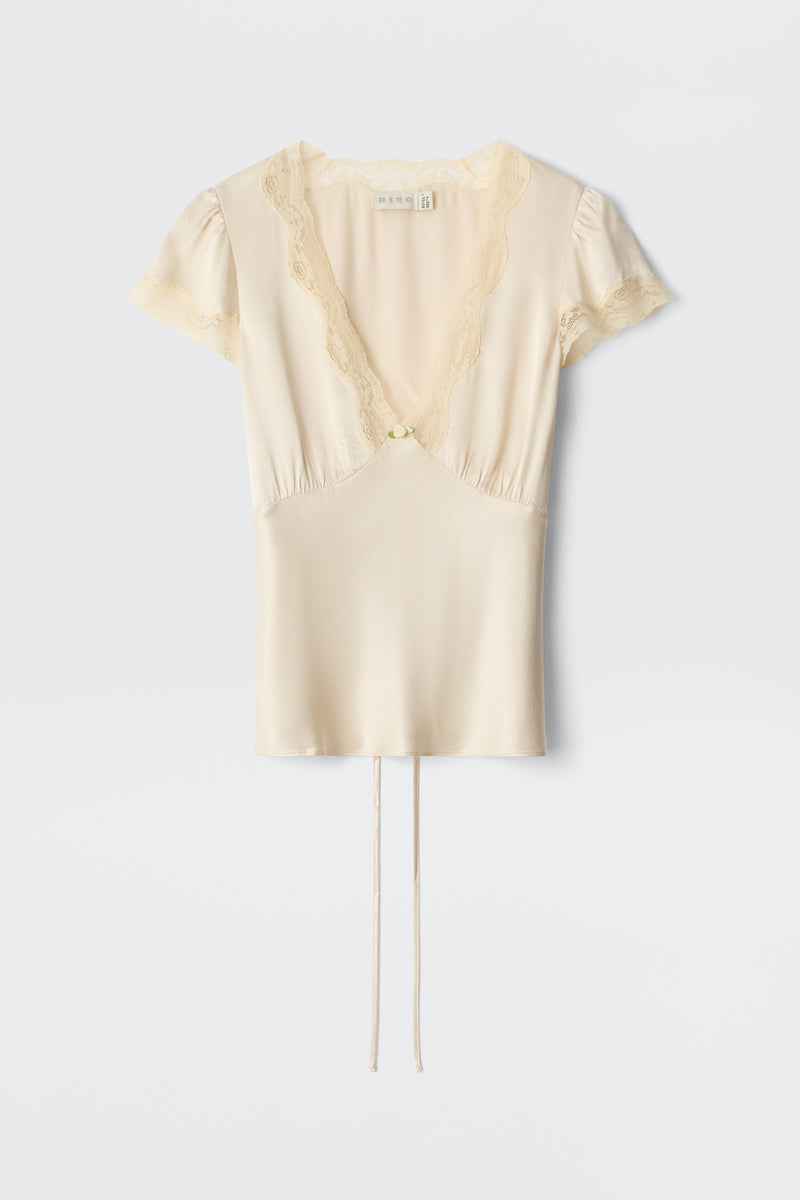 The Karia – Champagne is a cream-colored silk blouse featuring short sleeves, lace-trimmed deep V-neckline and sleeves, a center button, and two thin ties at the hem, shown against a plain white background.