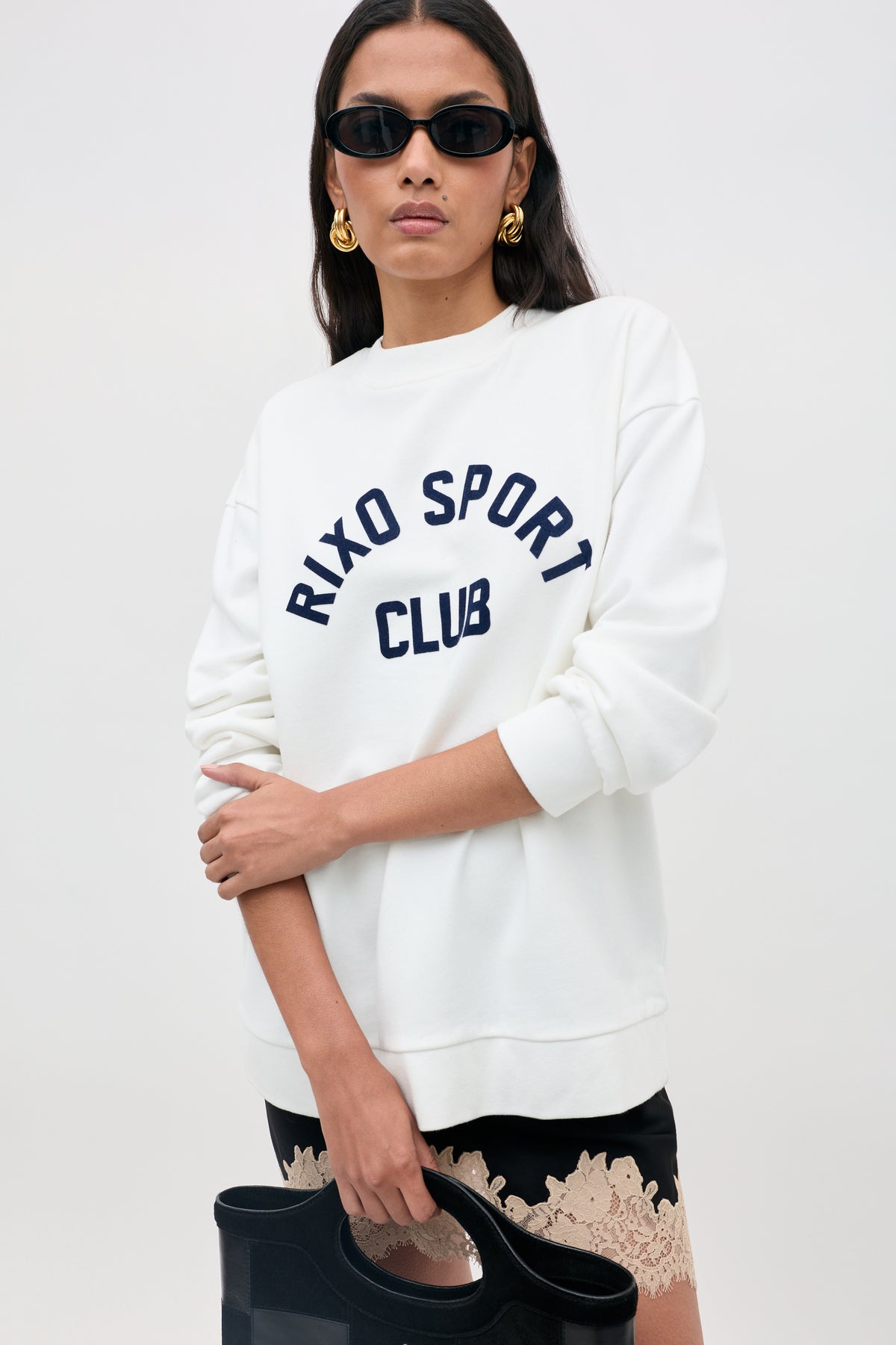 Woman wearing black sunglasses, gold earrings, and the Blaze - Rixo Sport Club White sweatshirt. She holds a black bag and wears a black skirt with beige lace trim. Plain light background.
