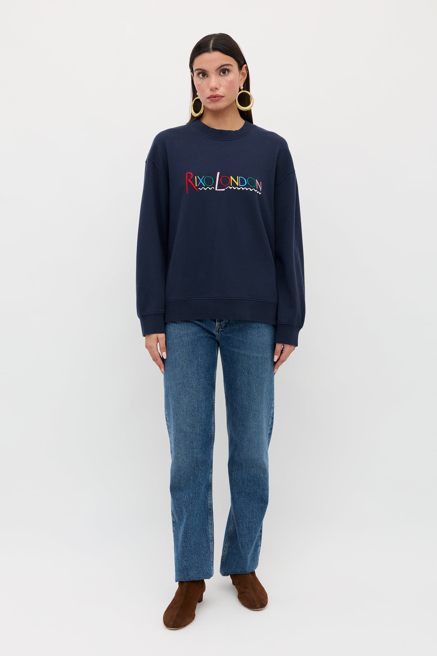 A woman stands against a plain white background wearing the Blaze - London Embroidery Navy sweatshirt from RIXO, paired with blue jeans, brown boots, and large hoop earrings. She faces forward with arms relaxed at her sides.