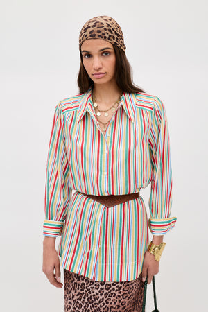 A person wearing the Ashlynn - Maxime Stripe Ivory shirt, a brown belt, layered gold necklaces, a gold bracelet, and a leopard print headscarf and skirt stands against a plain light background.