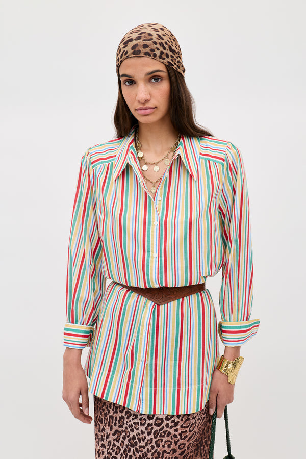 A person wearing the Ashlynn - Maxime Stripe Ivory shirt, a brown belt, layered gold necklaces, a gold bracelet, and a leopard print headscarf and skirt stands against a plain light background.