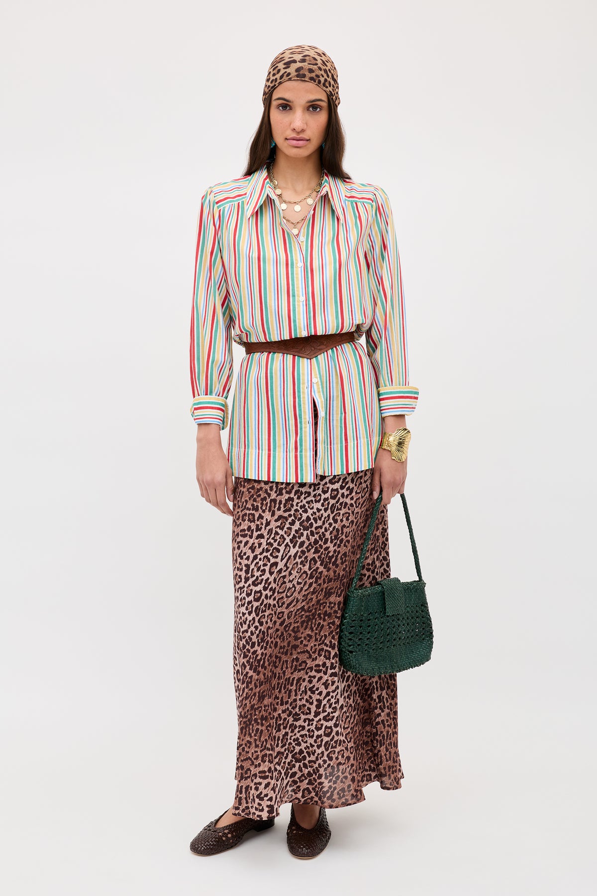 A woman in the Ashlynn - Maxime Stripe Ivory shirt, a leopard print maxi skirt, and matching headscarf stands with a dark green woven bag, layered necklaces, wide belt, and flat brown shoes against a plain light background.