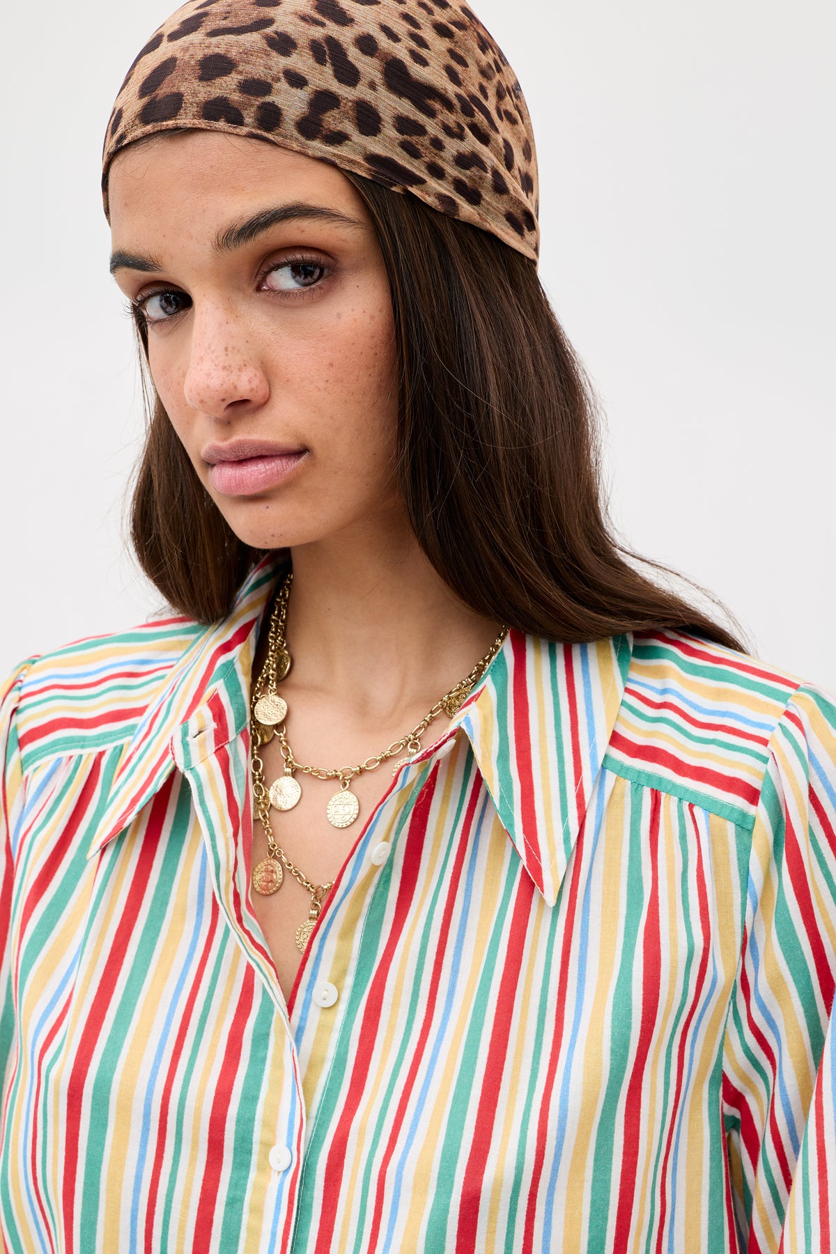 A woman with long brown hair wears a leopard-print headscarf, layered gold coin necklaces, and the Ashlynn - Maxime Stripe Ivory button-up shirt, looking at the camera against a plain background.