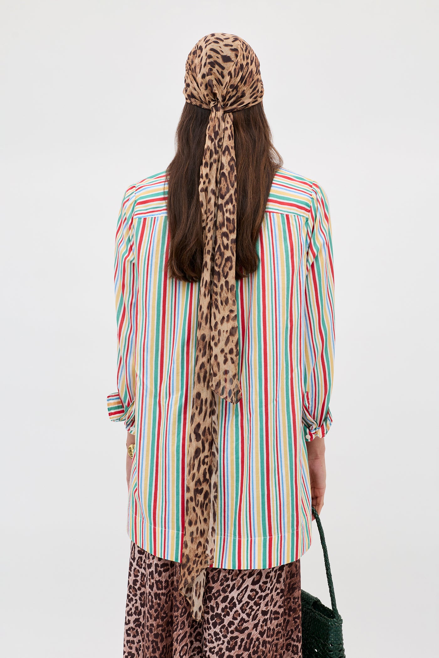 A person with long brown hair, seen from behind, wears the Ashlynn - Maxime Stripe Ivory shirt, a leopard print headscarf with tails down their back, a brown patterned skirt, and holds a green bag.