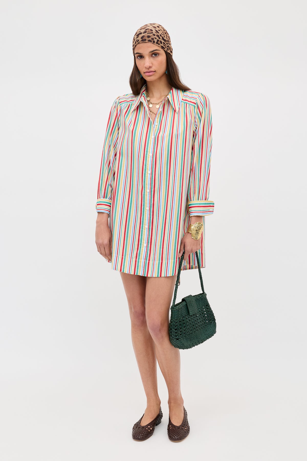 A woman stands against a white background wearing the Ashlynn - Maxime Stripe Ivory shirt dress, a brown headscarf, brown woven shoes, layered necklaces, and holding a green woven handbag.