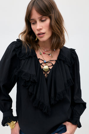 A woman wearing the Milia - Black ruffled blouse with lace details, styled with layered necklaces and wavy brown hair, looks down with a neutral expression against a light background.