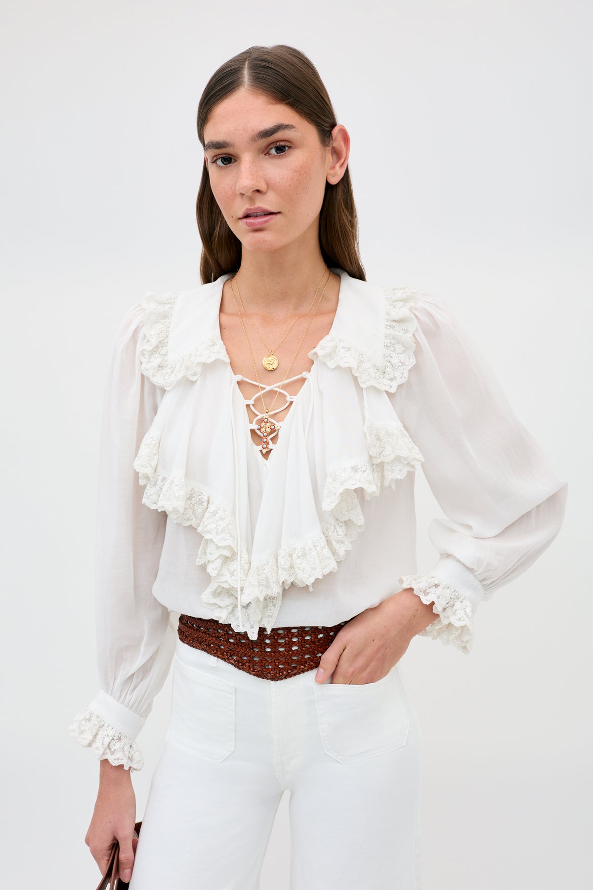 A woman with straight brown hair wears the Roxella - Tan belt over a white ruffled blouse and white pants. She accessorizes with gold necklaces and poses against a plain light background.