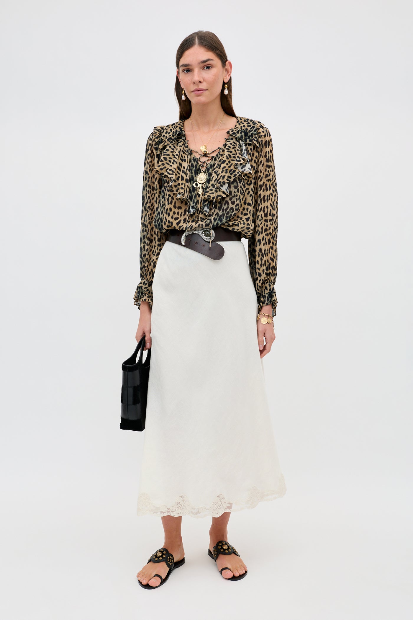 A woman with a neutral expression poses against a plain background wearing the Milia - Nina Leopard Brown blouse, a long white skirt, black sandals, gold jewelry, and carrying a black handbag.