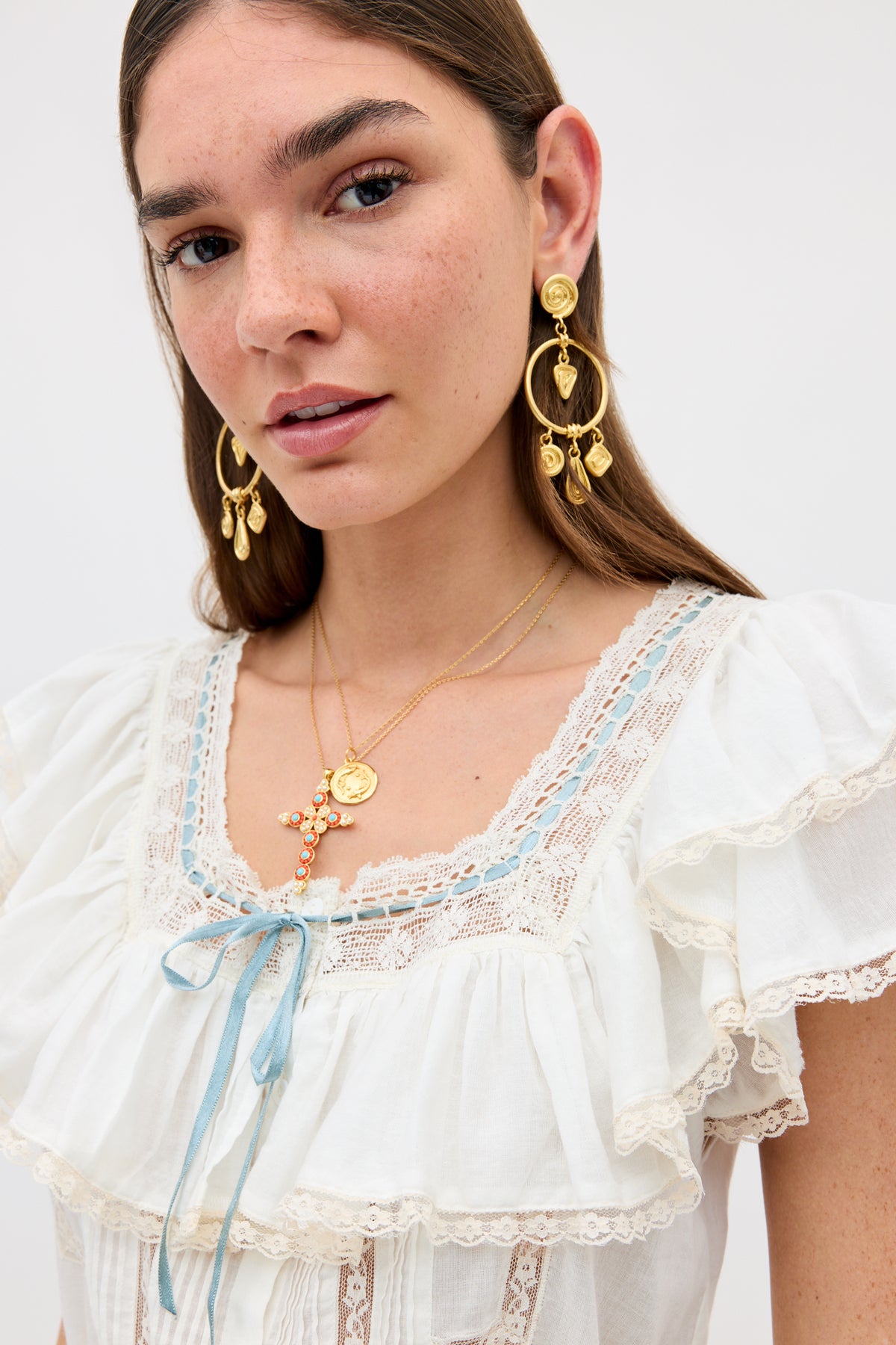 A woman with long brown hair and freckles wears the Melisa - Ivory blouse, featuring prairie-inspired white ruffles, lace details, and a blue ribbon, styled with large gold earrings and layered gold necklaces.