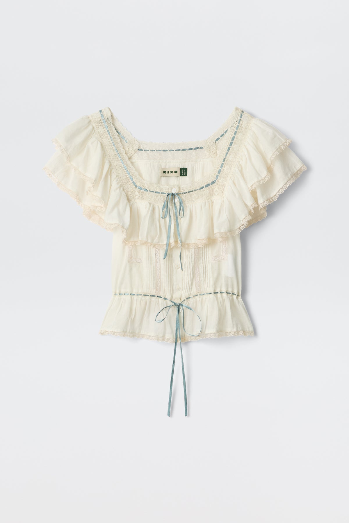 The Melisa - Ivory blouse features a prairie-inspired design with cream ruffles, lace trim, blue ribbon details, a gathered bust, and a waist tie, shown on a plain white background.