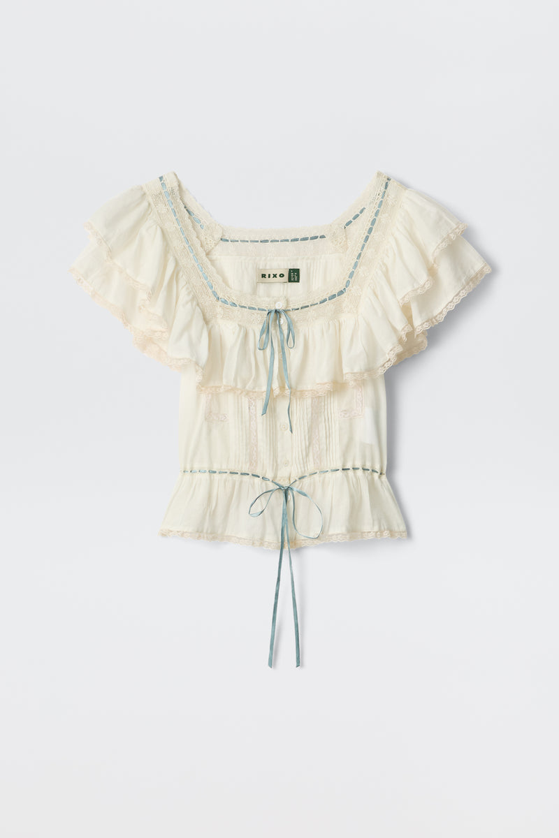 The Melisa - Ivory blouse features a prairie-inspired design with cream ruffles, lace trim, blue ribbon details, a gathered bust, and a waist tie, shown on a plain white background.