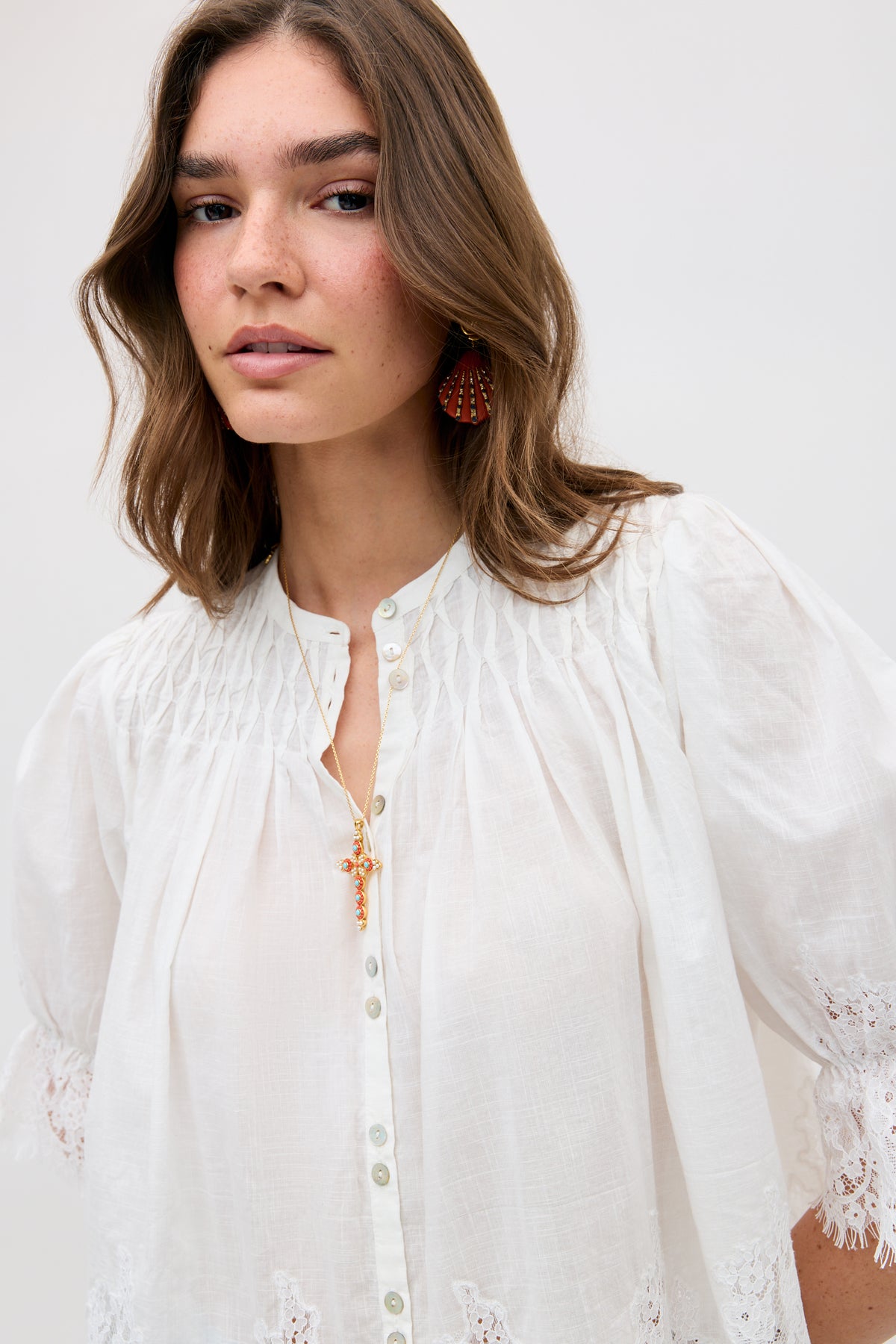 A woman with wavy brown hair wears the erika top ivory, a white button-up blouse with gathered details and lace trim. She pairs it with a gold pendant necklace and red statement earrings, standing against a plain light background.