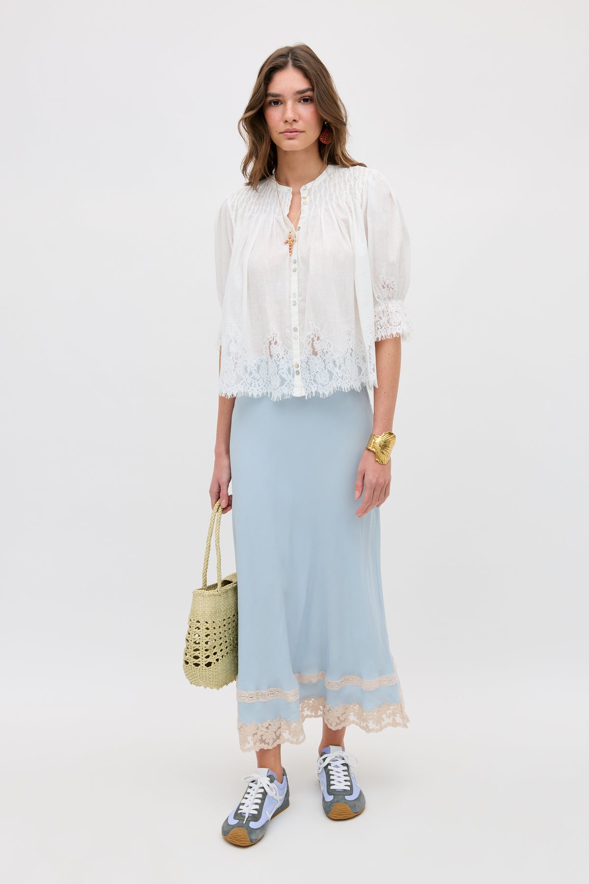A woman with loose hair and a neutral expression stands against a plain background wearing the erika top ivory, a light blue lace-hem maxi skirt, gray sneakers, and holding a woven straw bag.