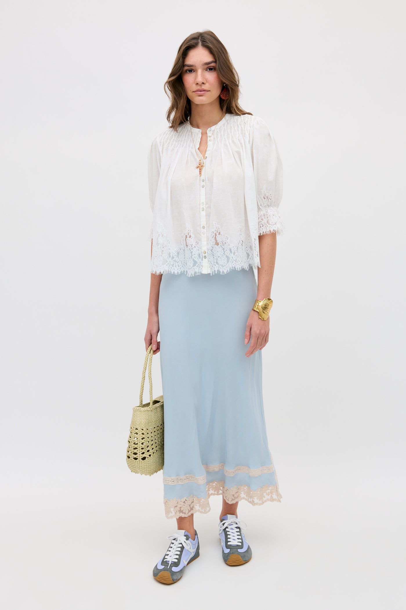 A woman with loose hair and a neutral expression stands against a plain background wearing the erika top ivory, a light blue lace-hem maxi skirt, gray sneakers, and holding a woven straw bag.