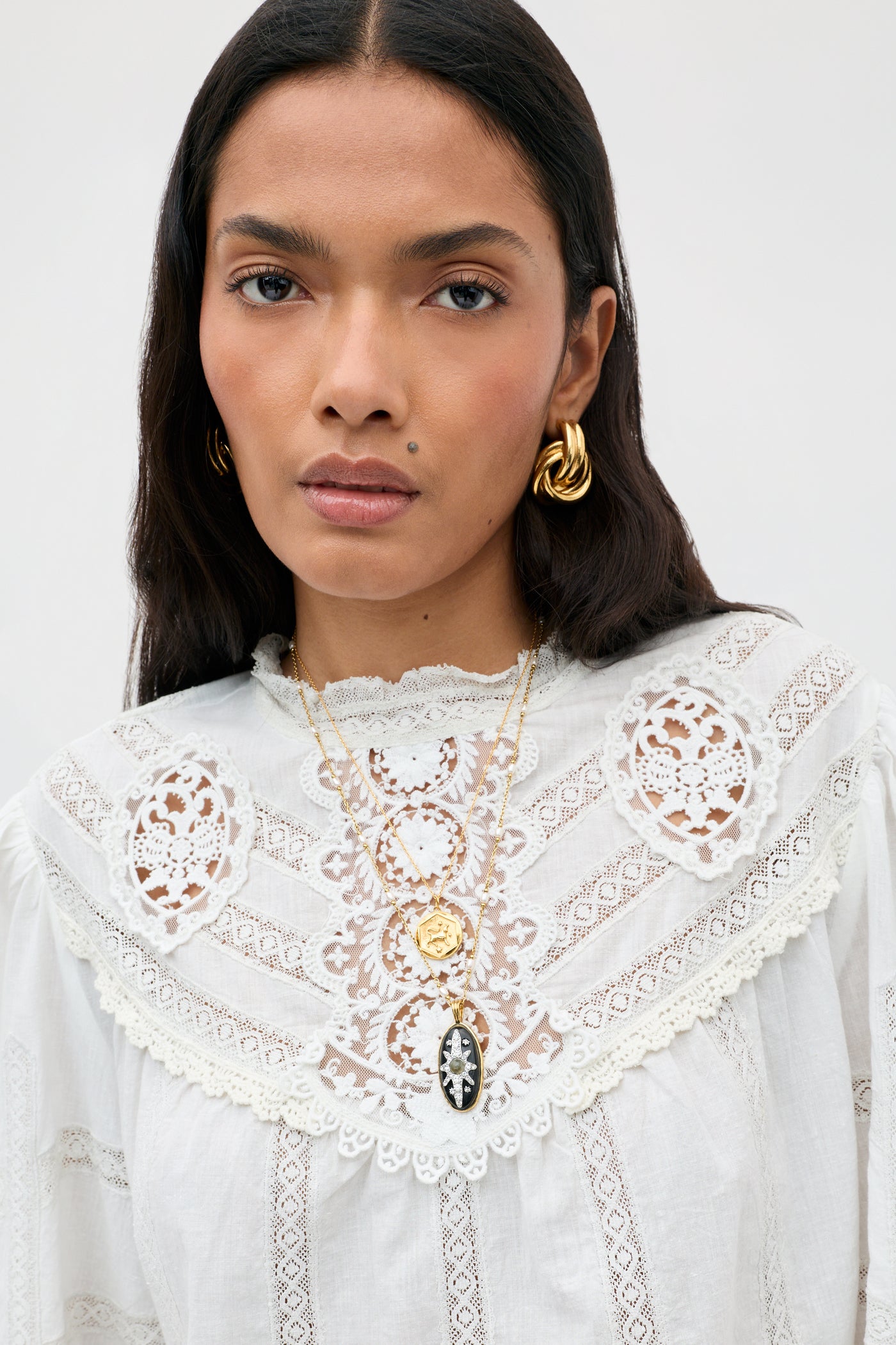 Wearing the Tamzin - Cream blouse, a woman with long dark hair pairs it with gold hoop earrings and layered gold pendant necklaces, standing against a plain light background.