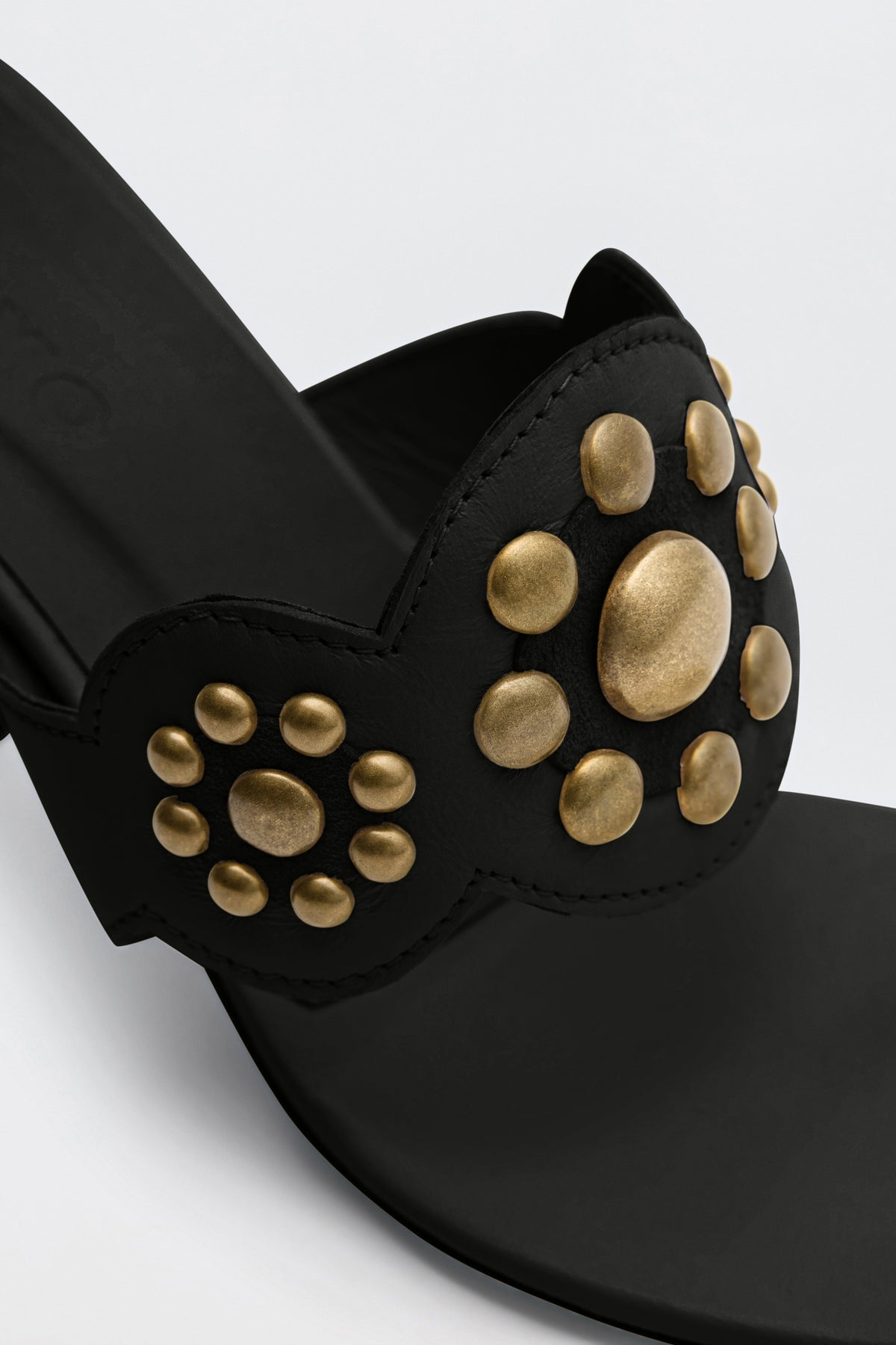 Close-up of the Esmeray - Black sandal, showcasing gold, rounded studs arranged in circular patterns on the front strap, set against a plain light background.