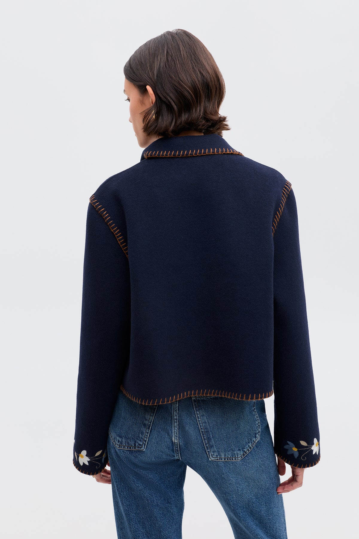 A person with short brown hair, seen from behind, wears the Sunday - Petal Embroidery Navy jacket, a luxurious navy wool blend with brown stitching and white floral embroidery near the cuffs, paired with blue jeans against a plain light background.